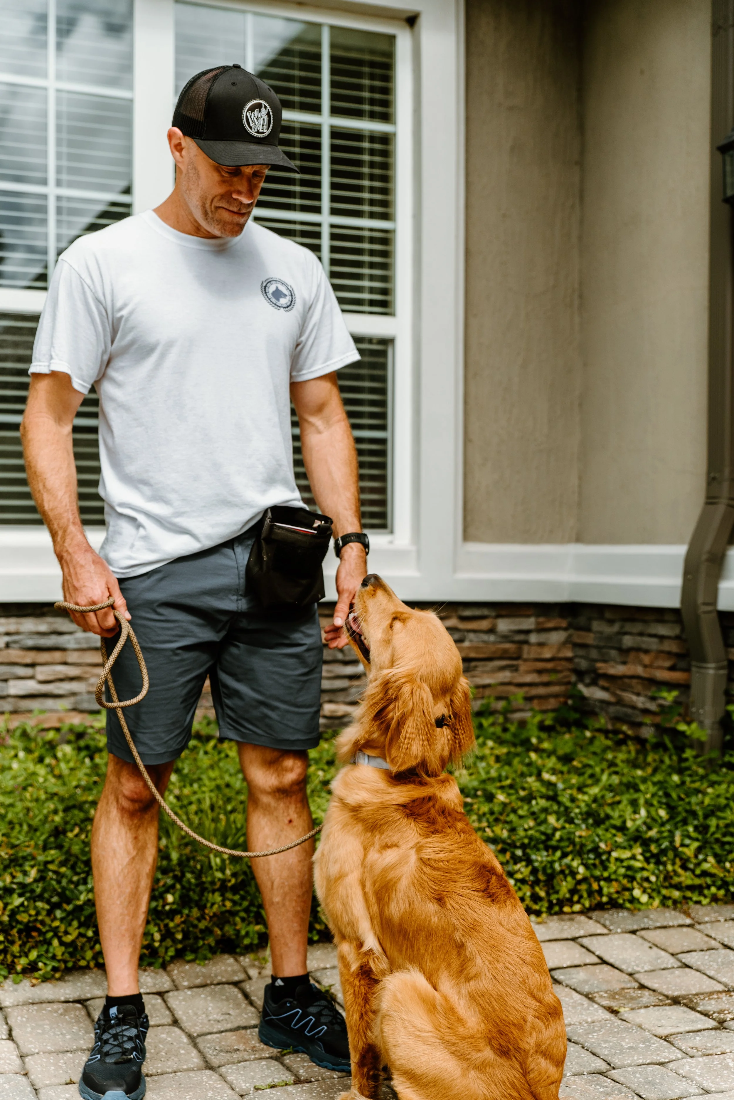 A man in a white t-shirt, shorts, and a cap training a golden retriever puppy outdoors on a stone patio, with a house and window in the background.