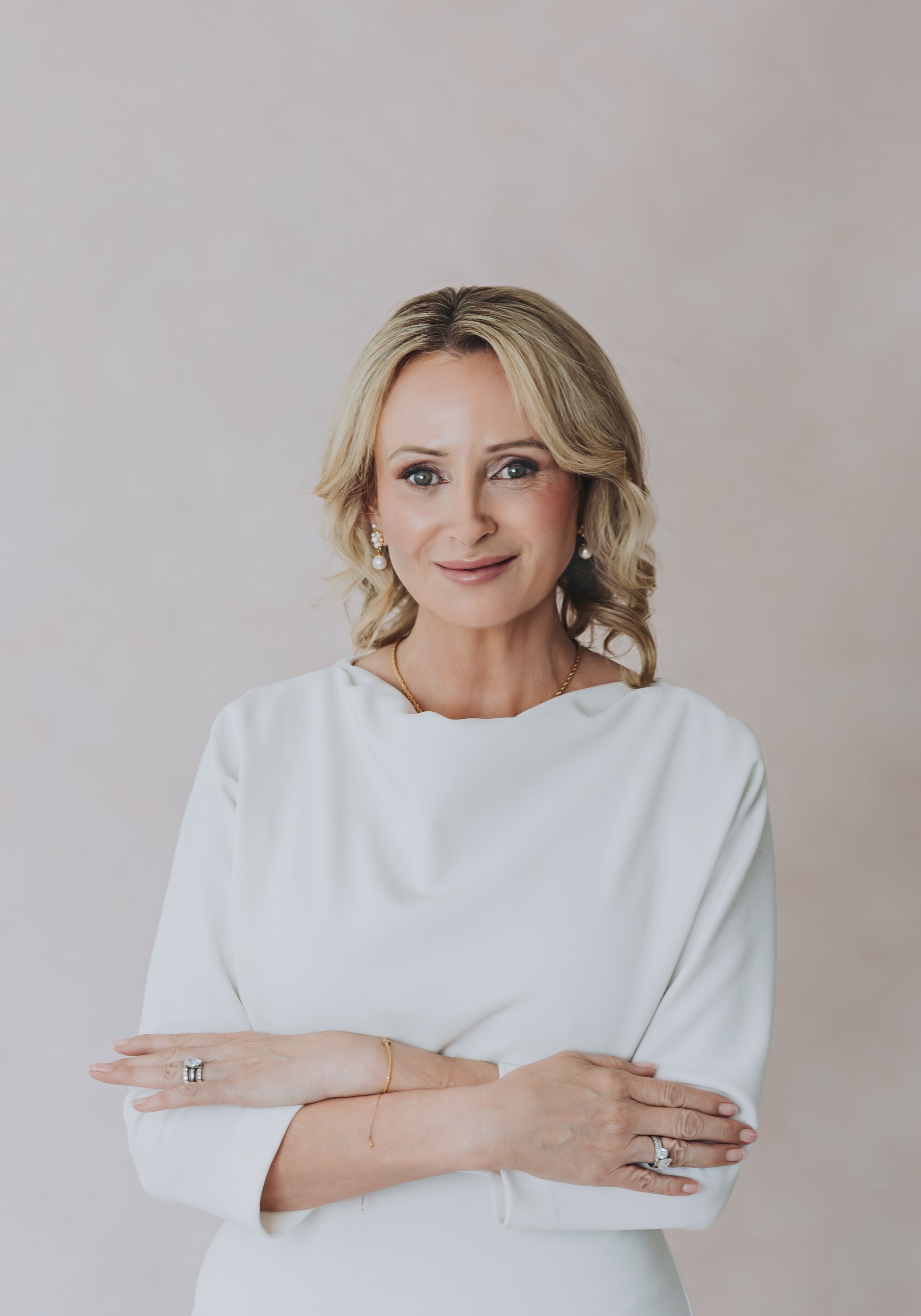 A woman with blonde hair styled in soft curls, wearing pearl earrings, a white dress, and jewelry, standing against a plain neutral background, smiling slightly with crossed arms.