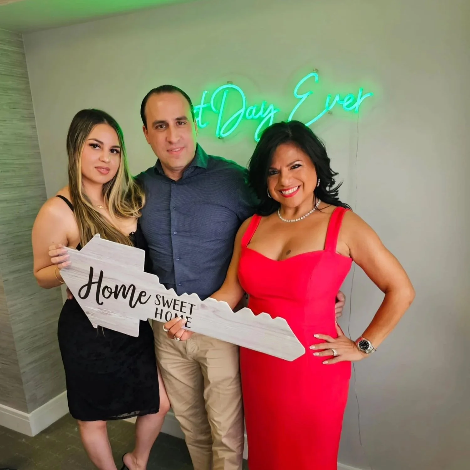 Three people standing together, holding a wooden sign shaped like the state of North Carolina that reads 'Home Sweet Home.' They are smiling, dressed nicely, with a neon sign behind them that says 'First Day Ever.'