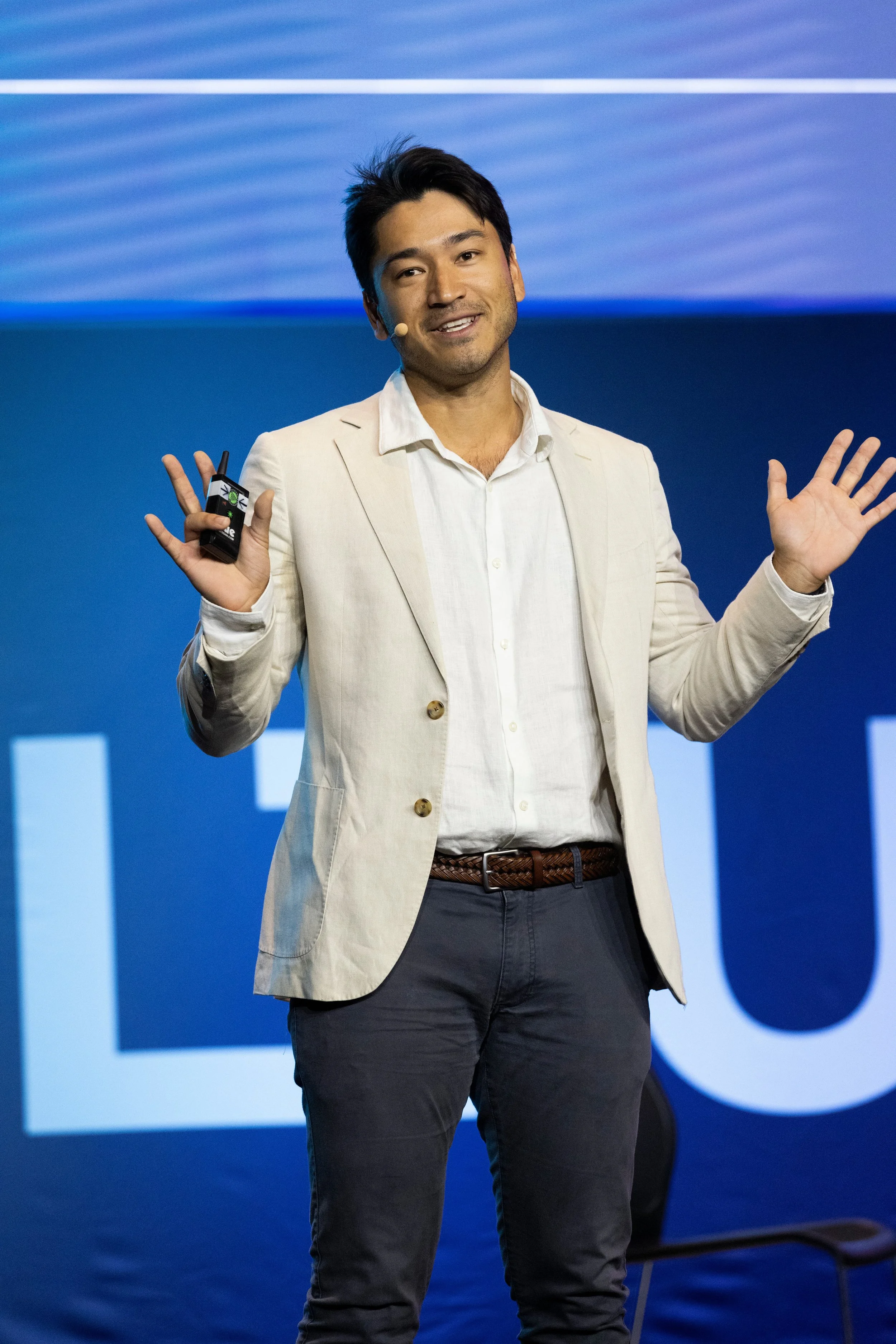 A man in a light-colored blazer and dark pants speaking on stage with a wireless microphone headset, gesturing with one hand, in front of a blue background with large text.