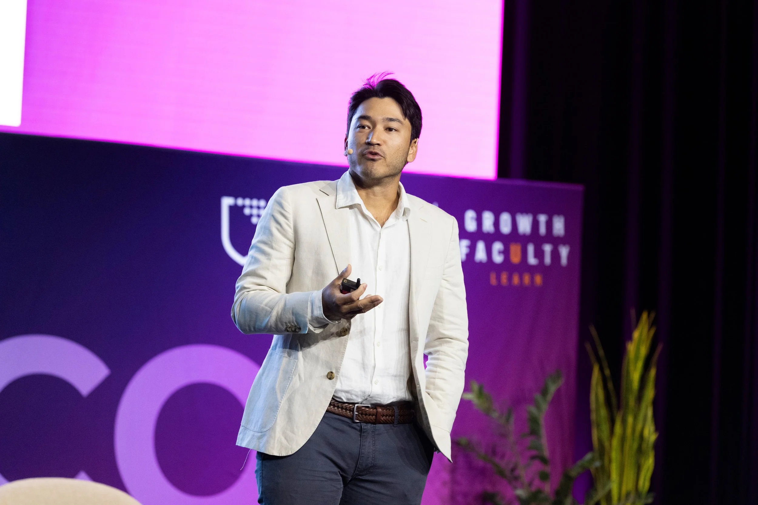 A man in a light-colored blazer giving a presentation on stage with a large pink and purple screen behind him.