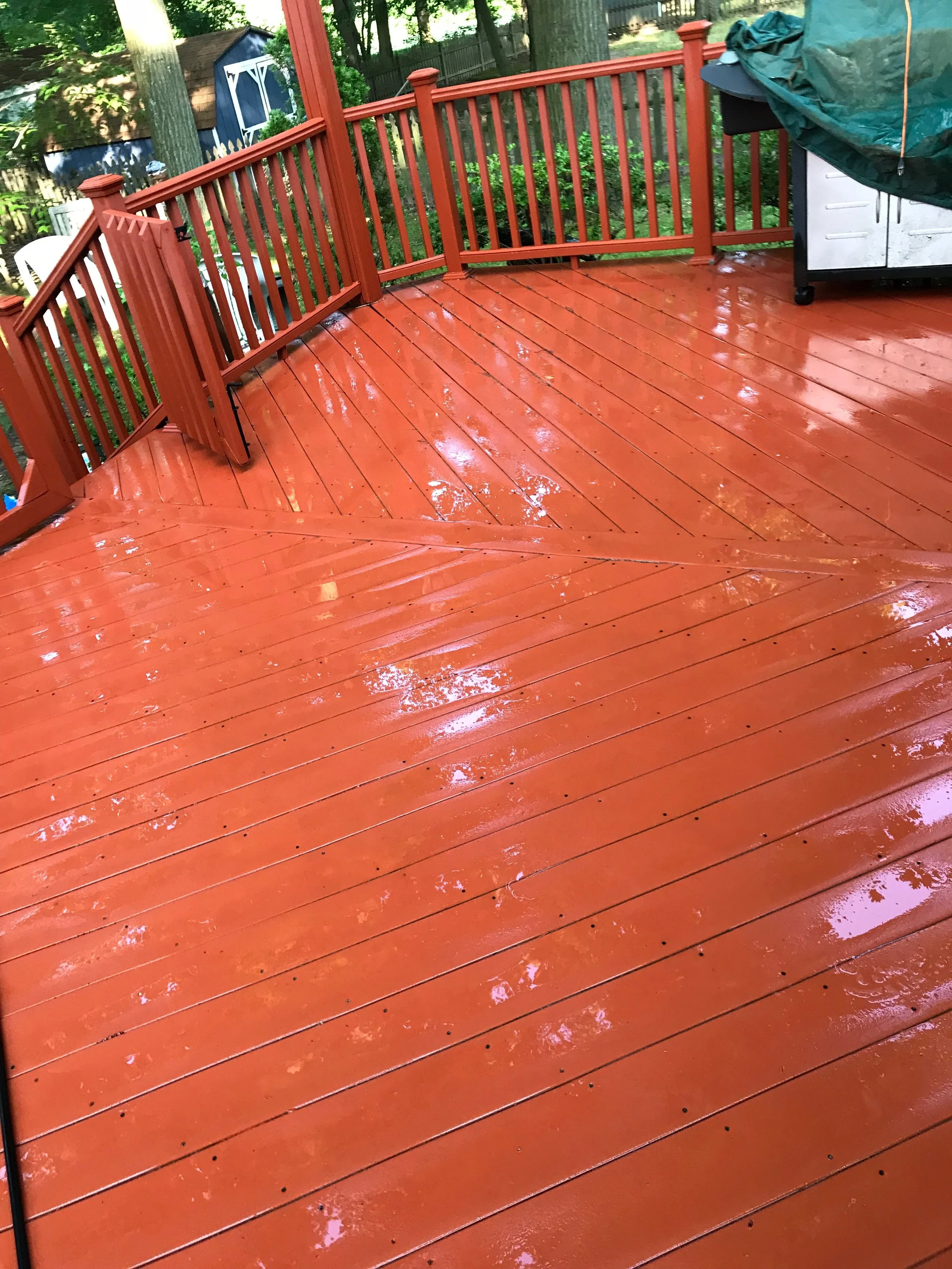 A wet red wooden deck with reflections of trees on its surface, surrounded by a wooden railing, with garden tools and a barbecue grill in the background.