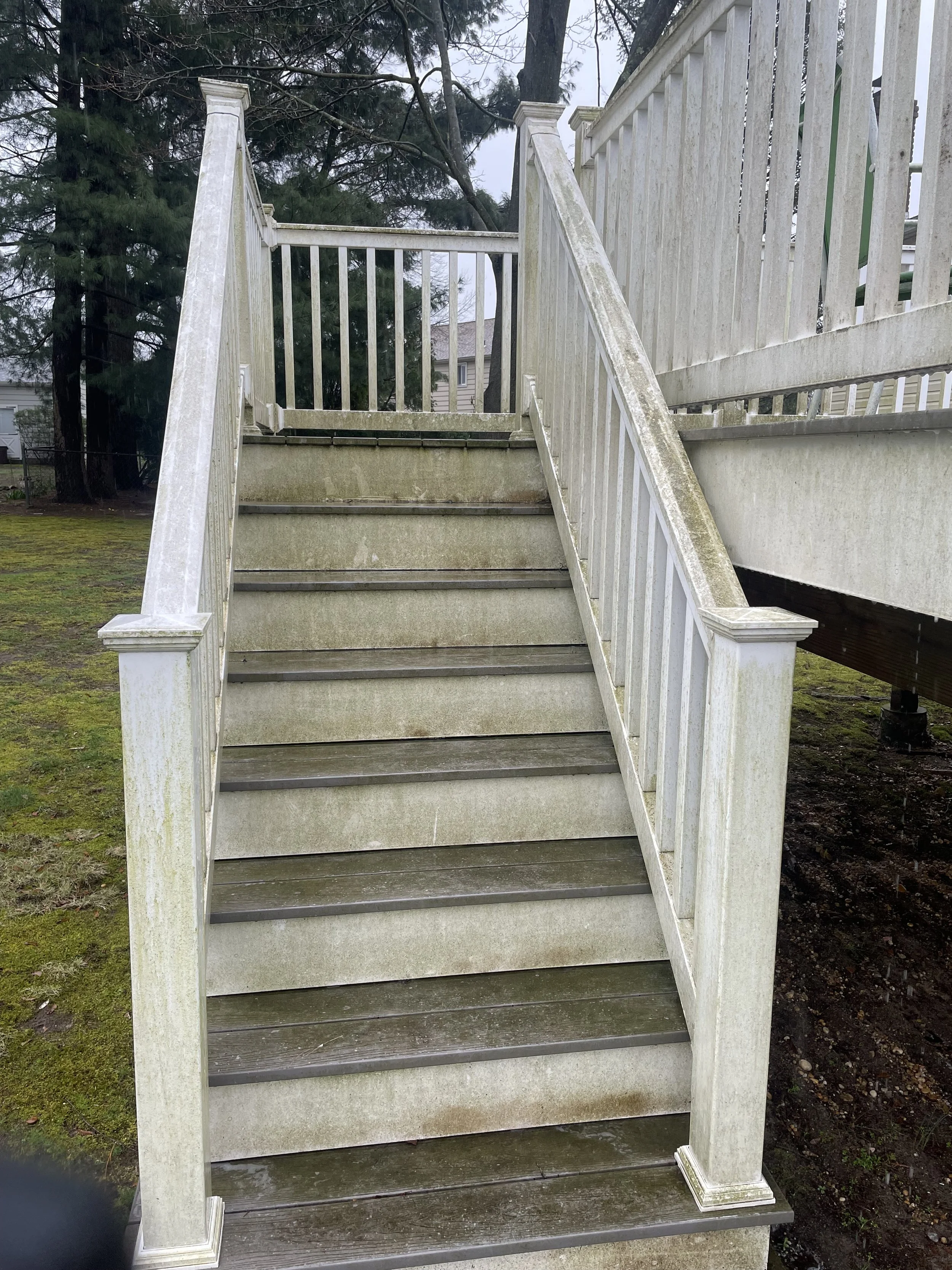 Outdoor wooden staircase with white railing leading to an elevated deck, weathered and mossy, with trees and a cloudy sky in the background.