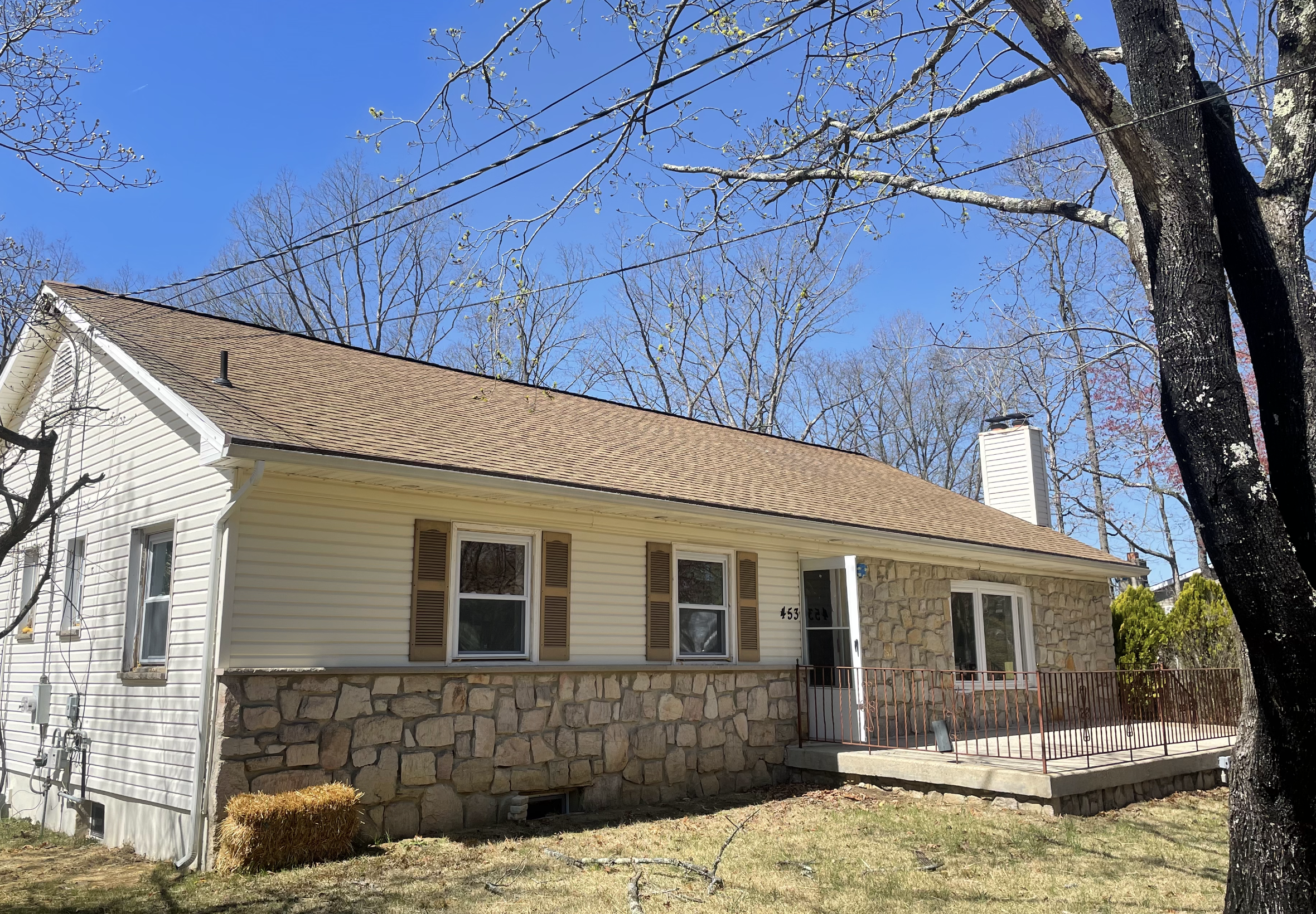 A single-story house with a stone foundation, cream siding, and brown shutters, with a small porch and a fenced deck, under a clear blue sky.