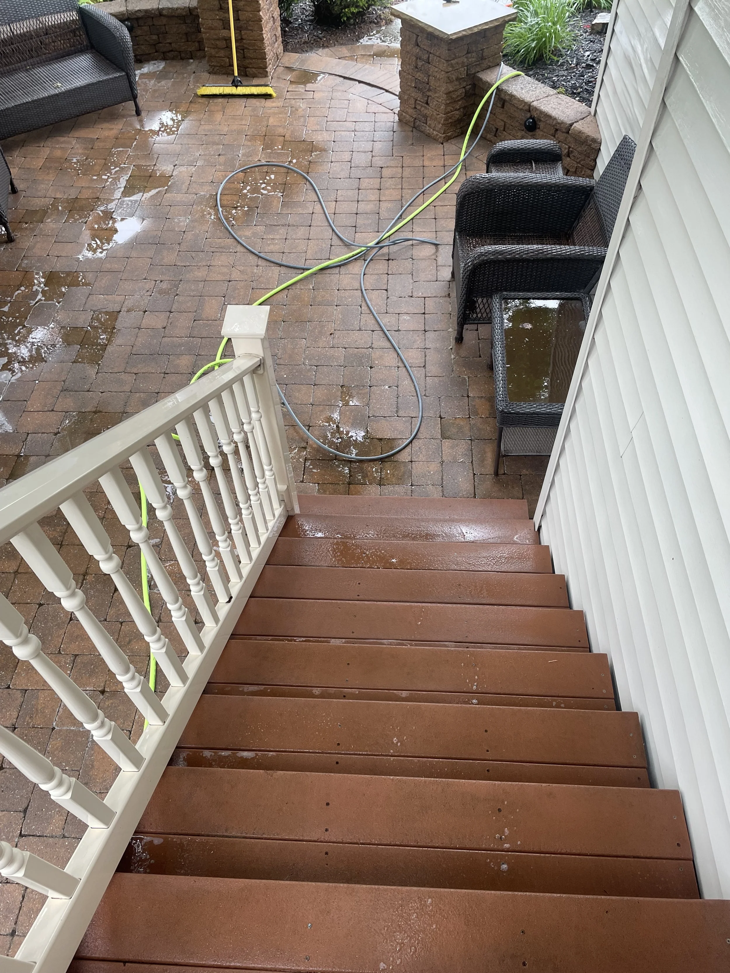 Wet brick patio with garden hose, yellow broom, black outdoor furniture, and a white staircase leading down to the patio.