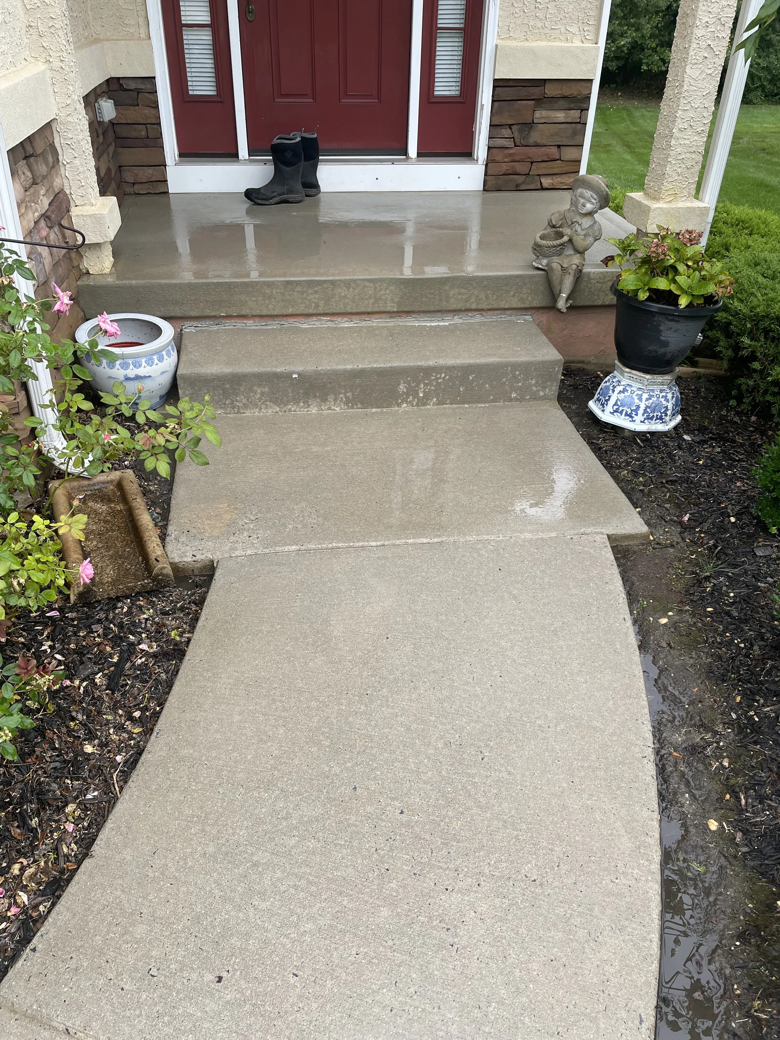 Concrete front porch with red door and black boots, potted plants, a small statue of a girl, and wet concrete walkway.