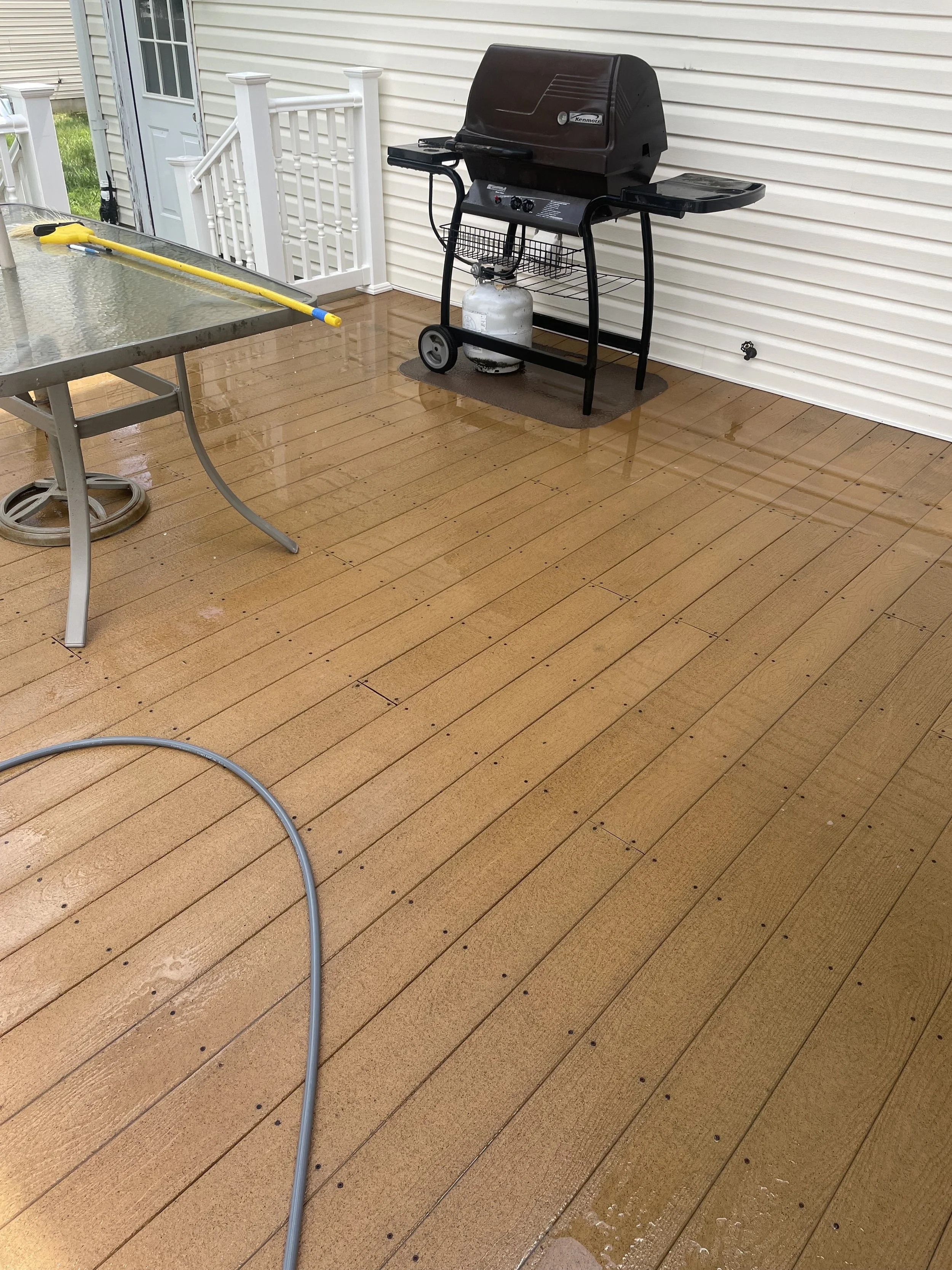 Clean, wet wooden deck with a gas barbecue grill and a glass-top outdoor table with a yellow cleaning tool resting on it.