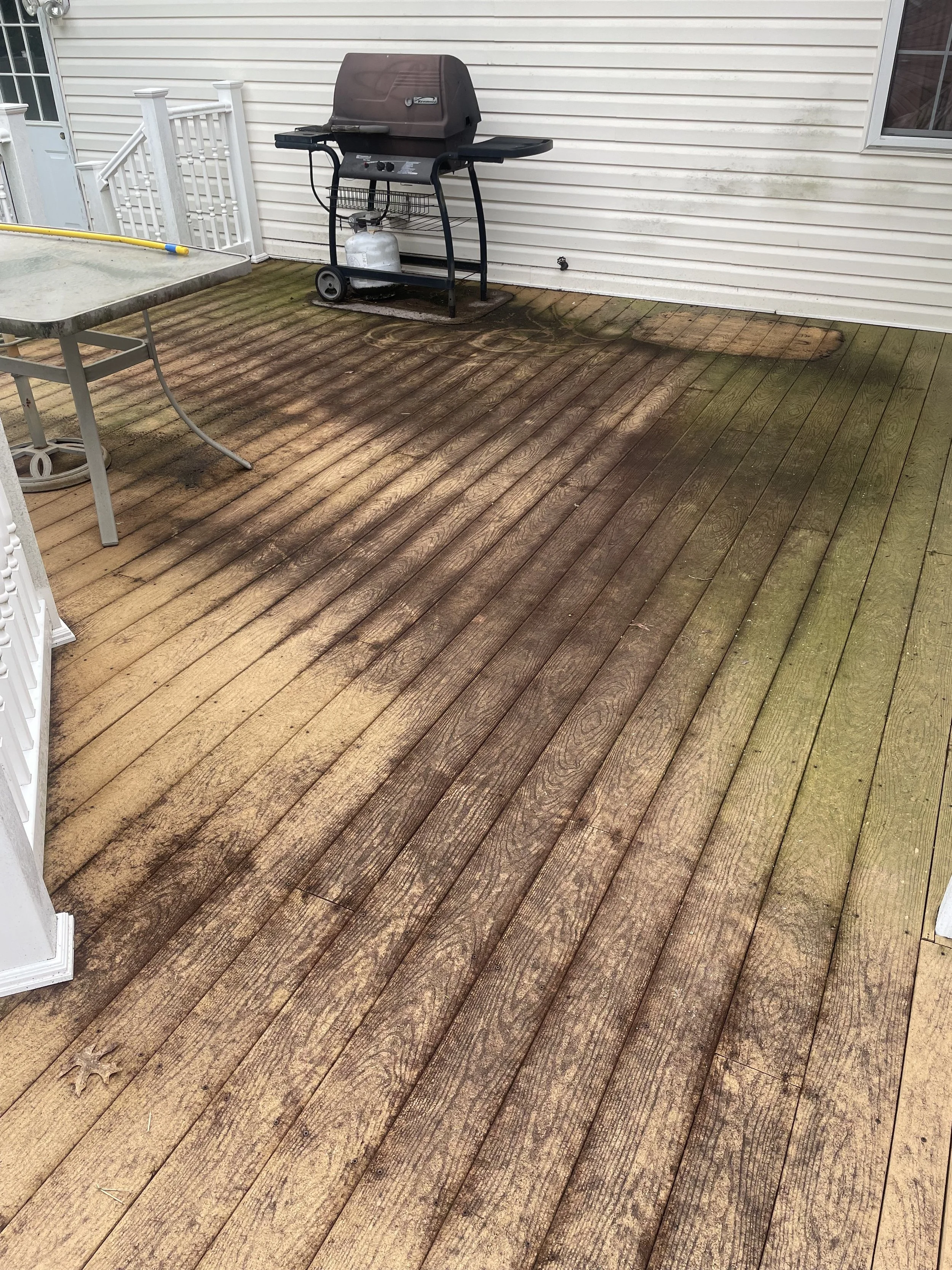 Dirty and mossy wooden deck with a grill and a table on it, next to a house with white siding.