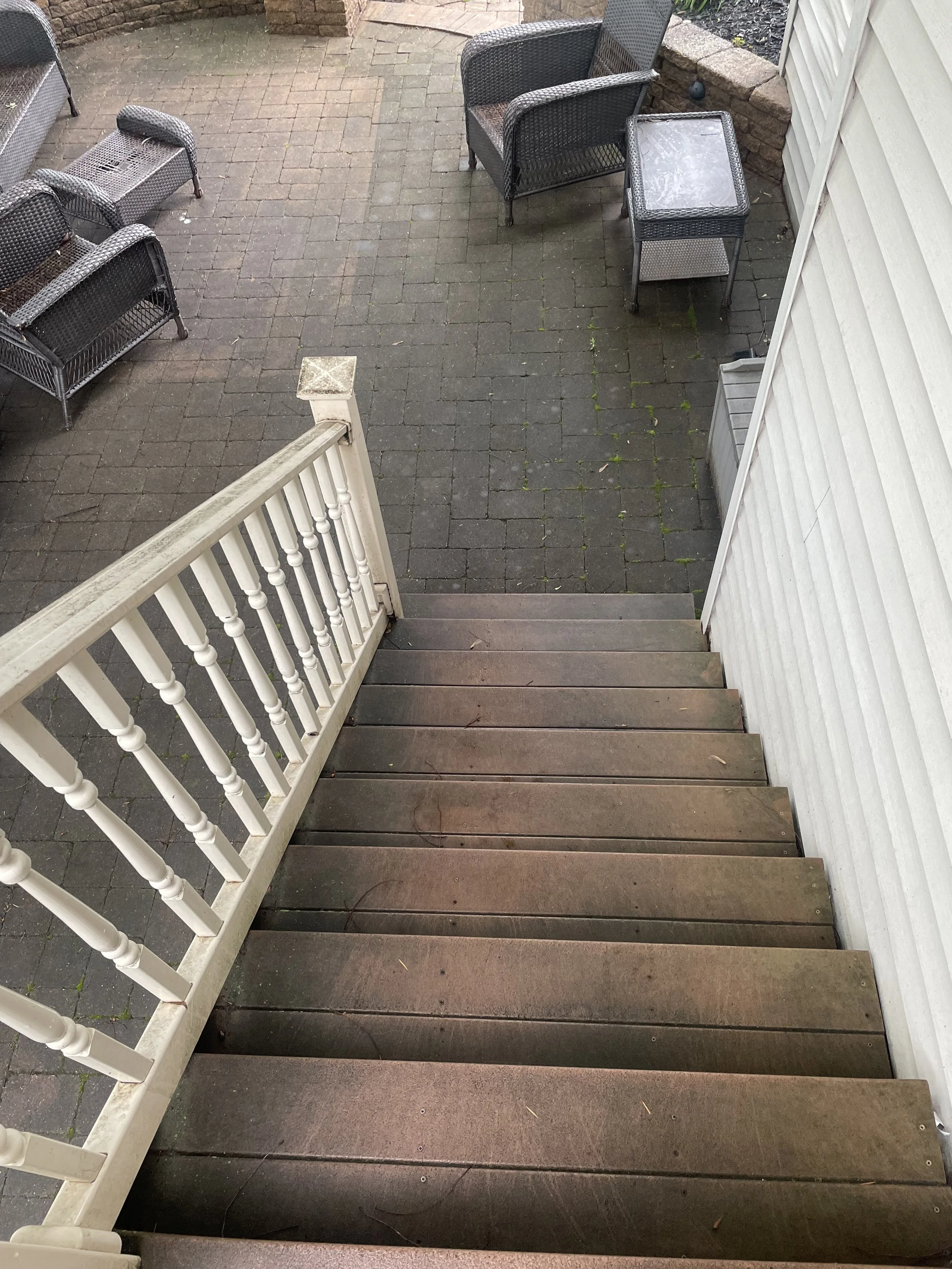 View from top of wooden stairs looking down onto an outdoor patio with patio furniture, including several wicker chairs and a small table, surrounded by a brick retaining wall and white siding.