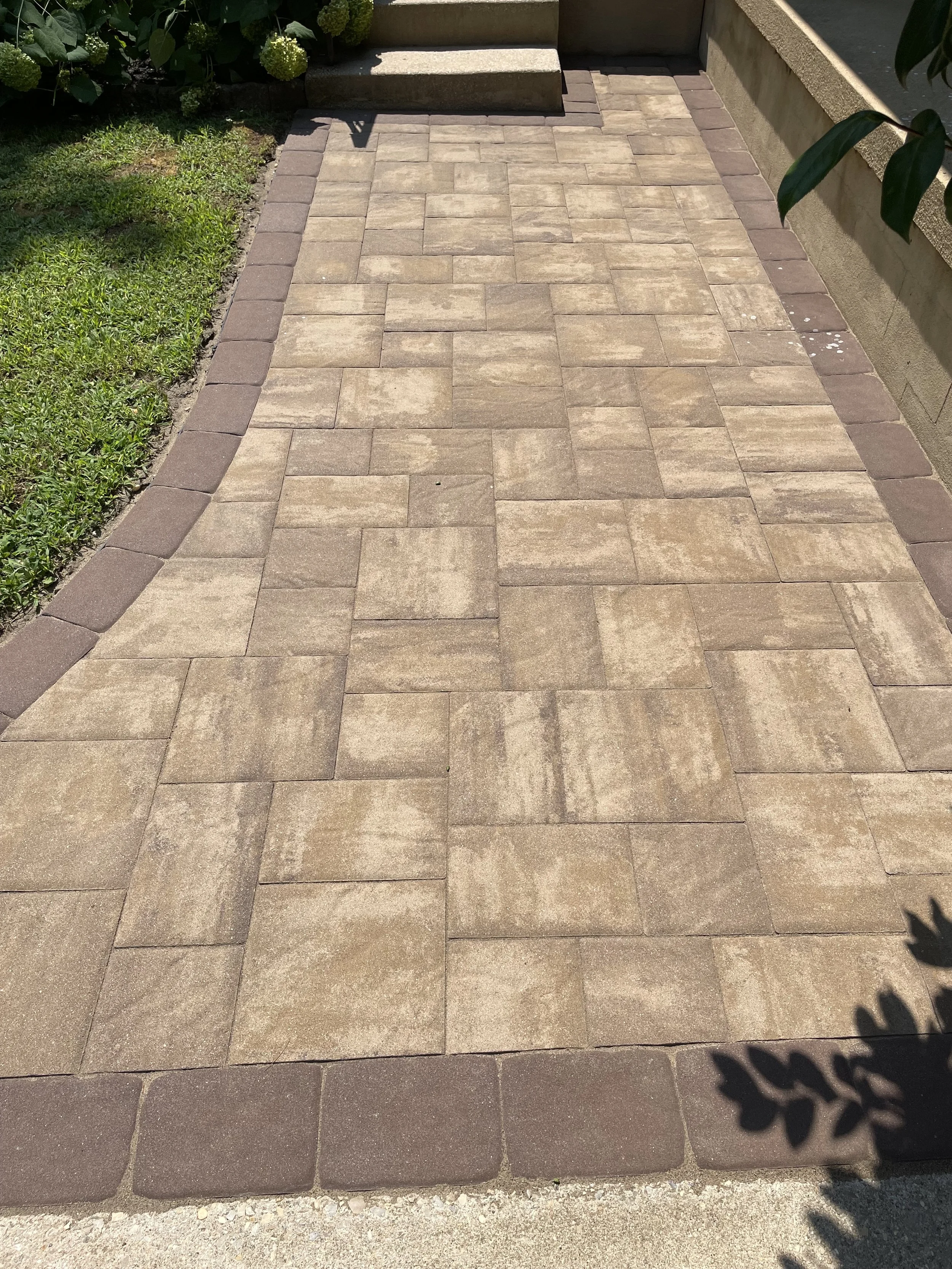A concrete patio with beige pavers bordered by dark red bricks, with stairs and greenery in the background.
