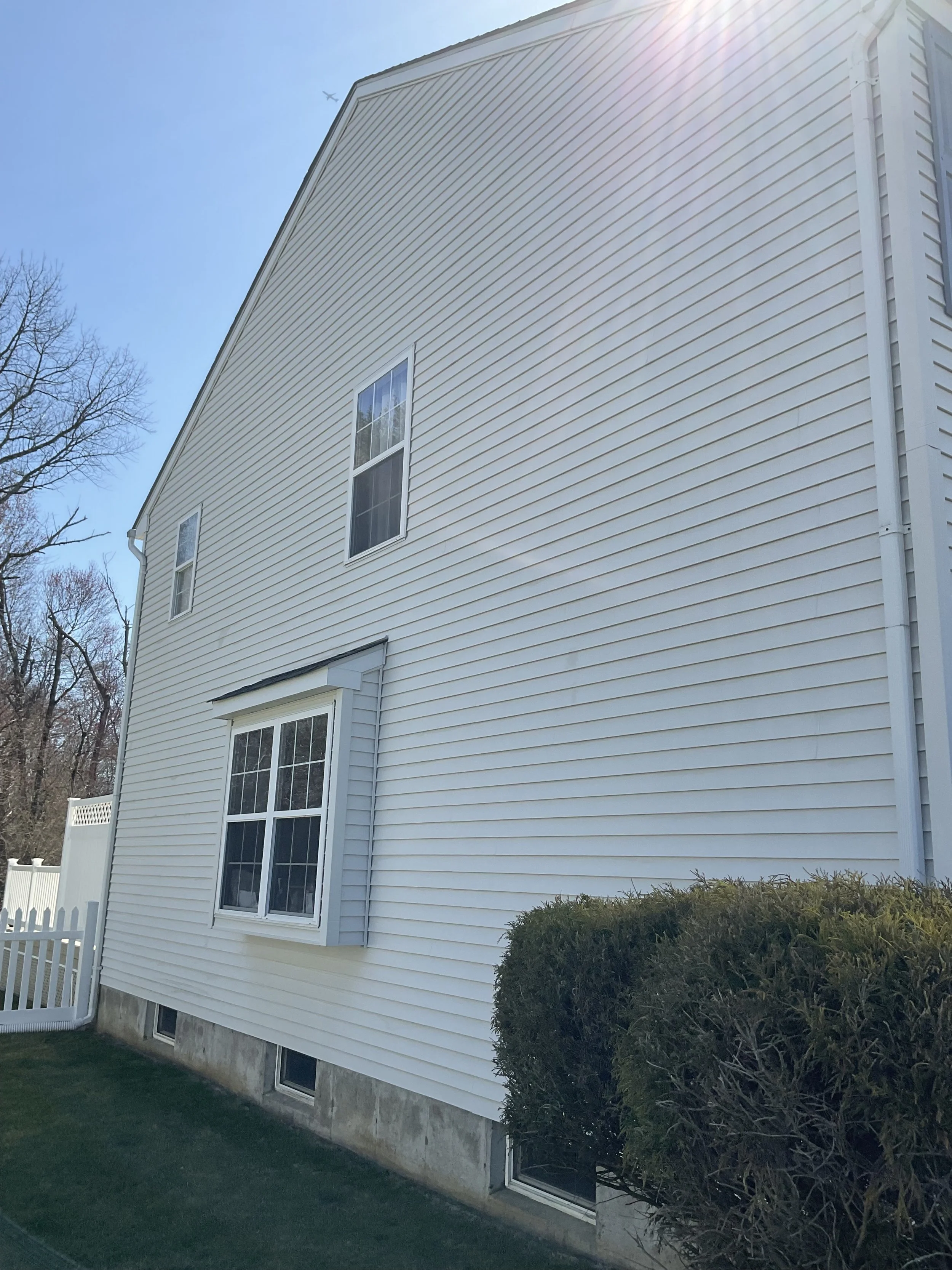 Side view of a white house with vinyl siding, two large windows with decorative grids, a small window at the basement level, and a bush in front. Clear blue sky with sunlight visible.