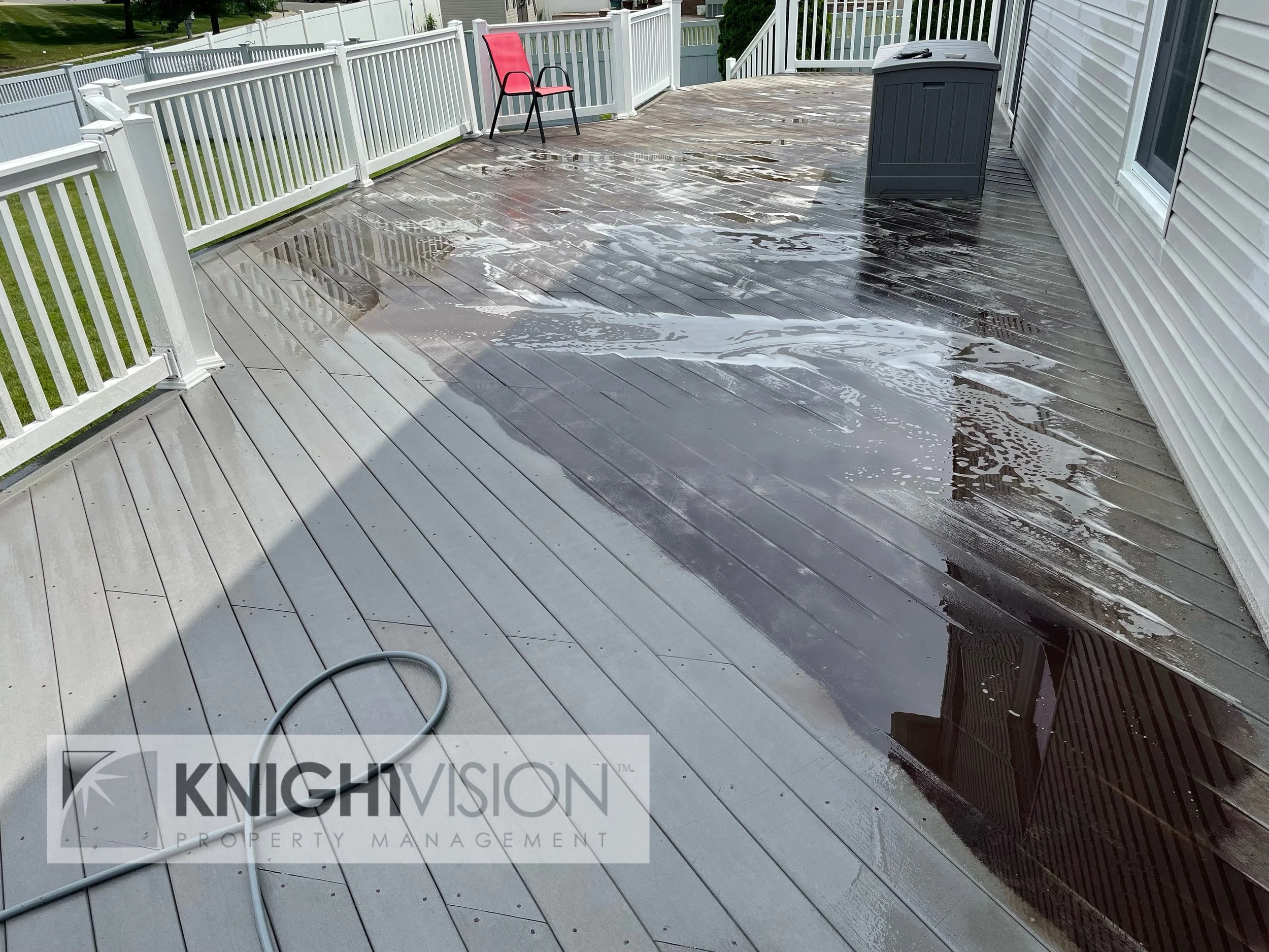 Wet wooden deck on an outdoor patio with a red chair, a gray storage box, and a white railing. A garden hose lies on the deck, and water is visible on the surface.