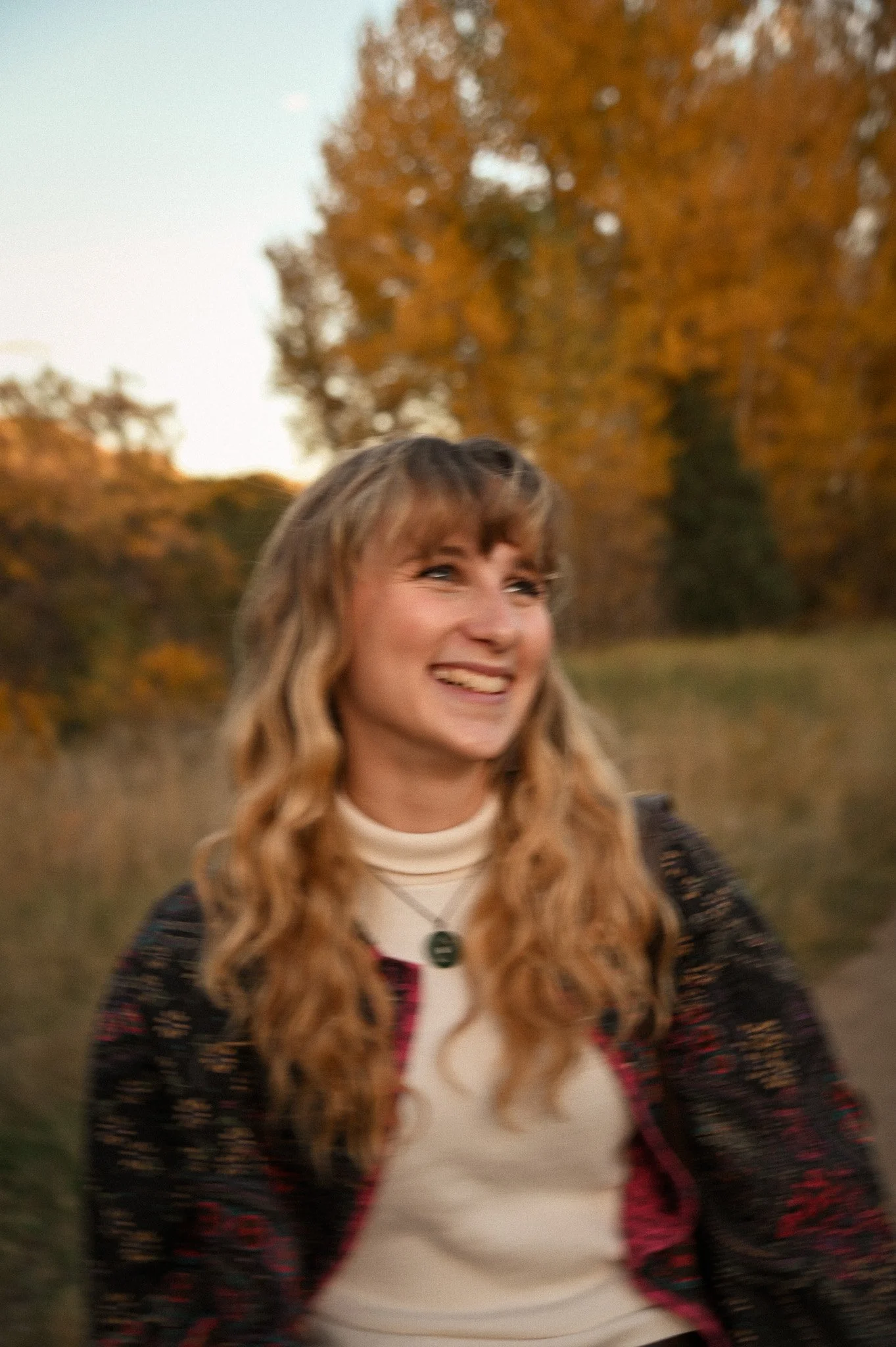 A young woman with long wavy blonde hair, smiling, outdoors with autumn-colored trees in the background.