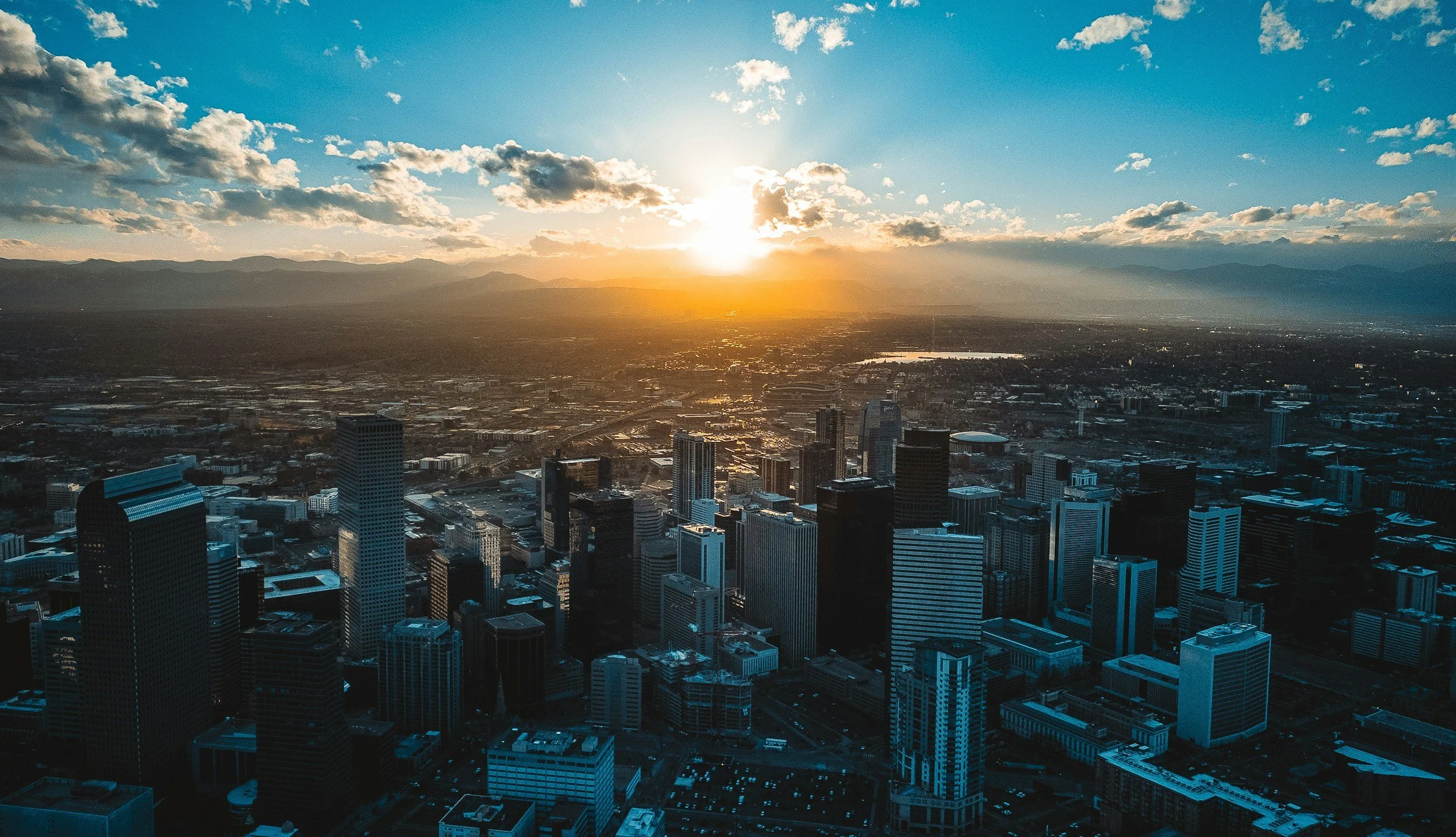 Aerial view of a city skyline at sunset with tall buildings in the foreground and mountains in the background.