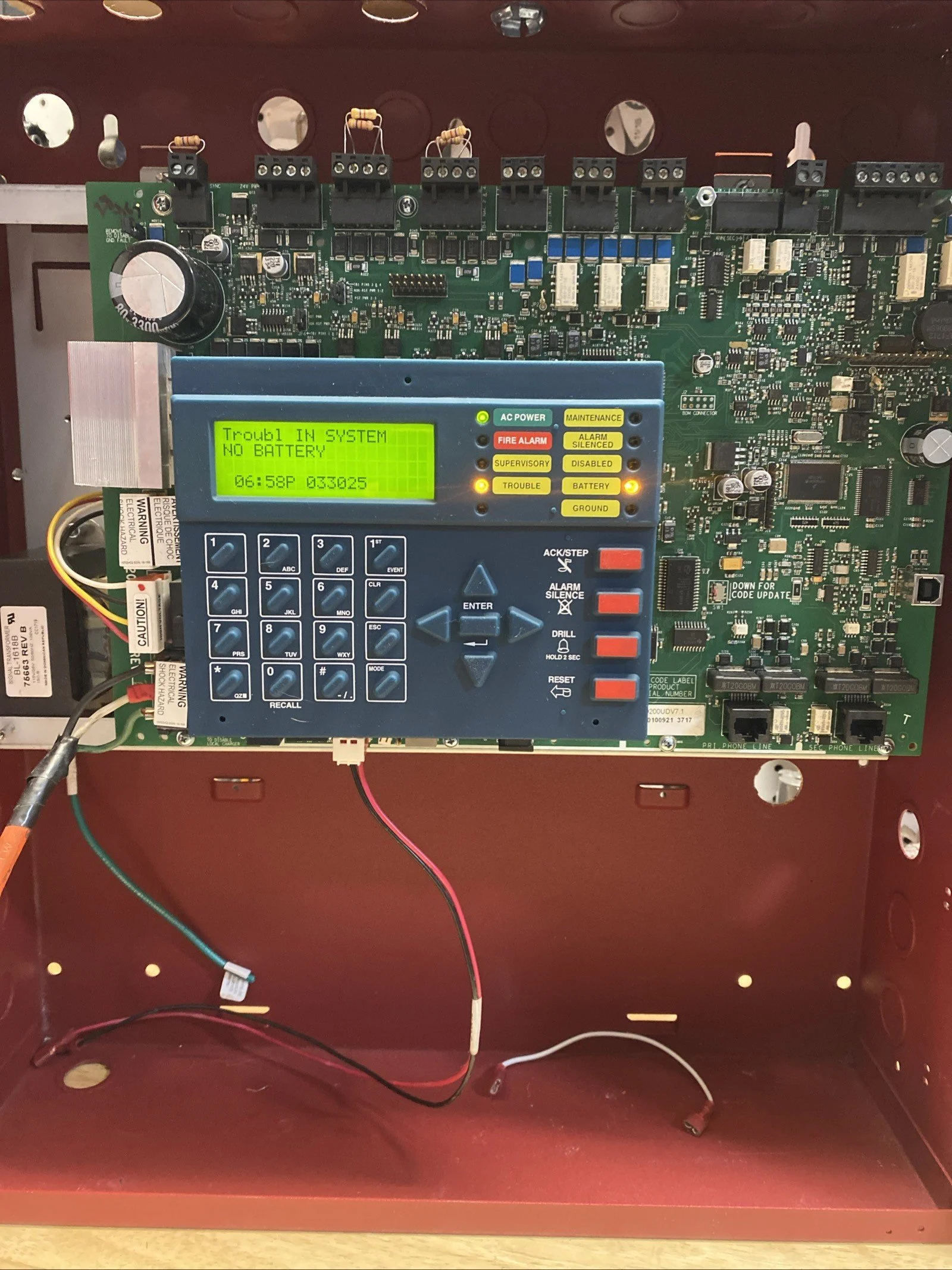 Interior of a security alarm control panel with a green circuit board and a blue keypad display showing 'Trouble IN SYSTEM, NO BATTERY,' and an additional message with date and time. The panel has various wires connected and is mounted inside a red metal box.