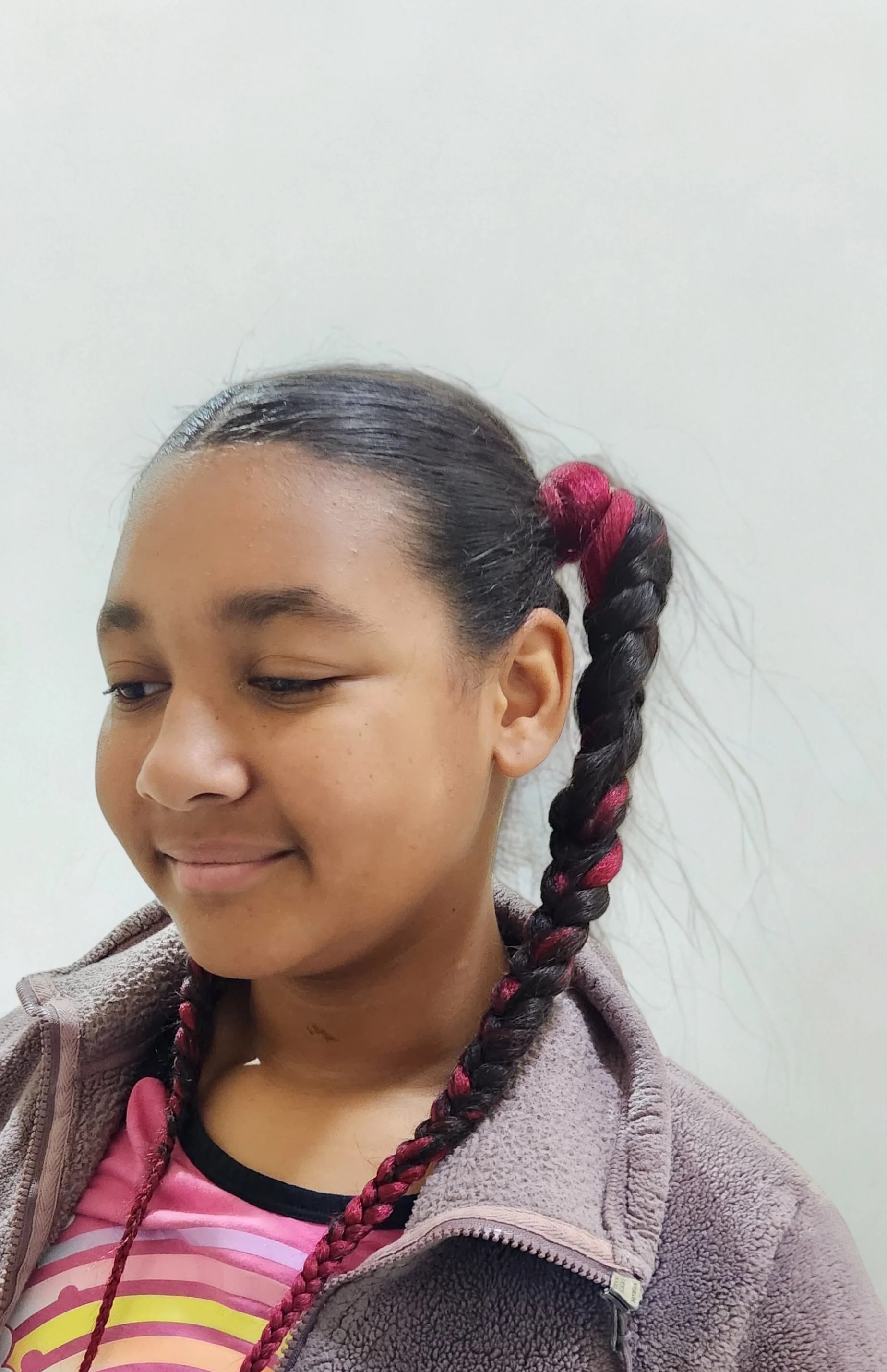 A young girl with dark hair styled in a long braid with red highlights, wearing a pink and yellow shirt and a brown fleece jacket.