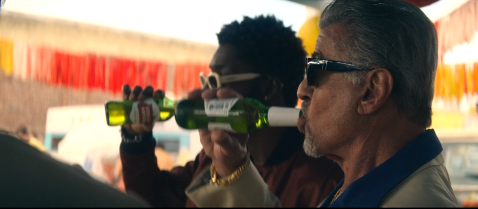 Two men standing outdoors, one wearing sunglasses and a beige jacket drinking from a green glass bottle, and the other holding a similar bottle near his face, with strings of red and yellow decorations in the background.