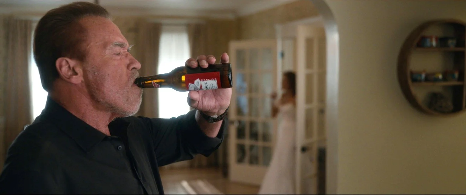 An older man with gray hair and beard wearing a black shirt drinks beer from a bottle inside a house, with a woman in a white dress in the background