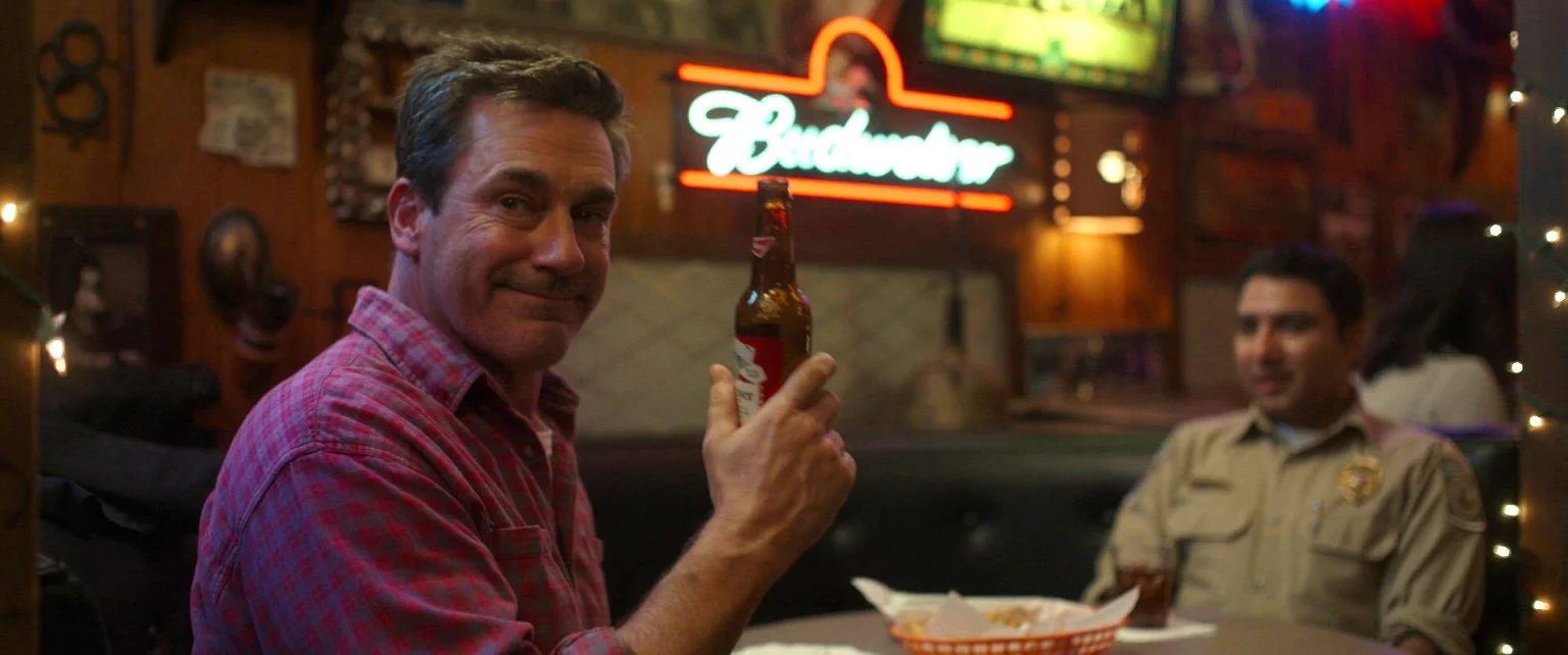 A man in a red plaid shirt holding a Budweiser beer bottle in a bar or restaurant setting, with a neon Budweiser sign in the background and a woman in a sheriff's uniform sitting at the table.