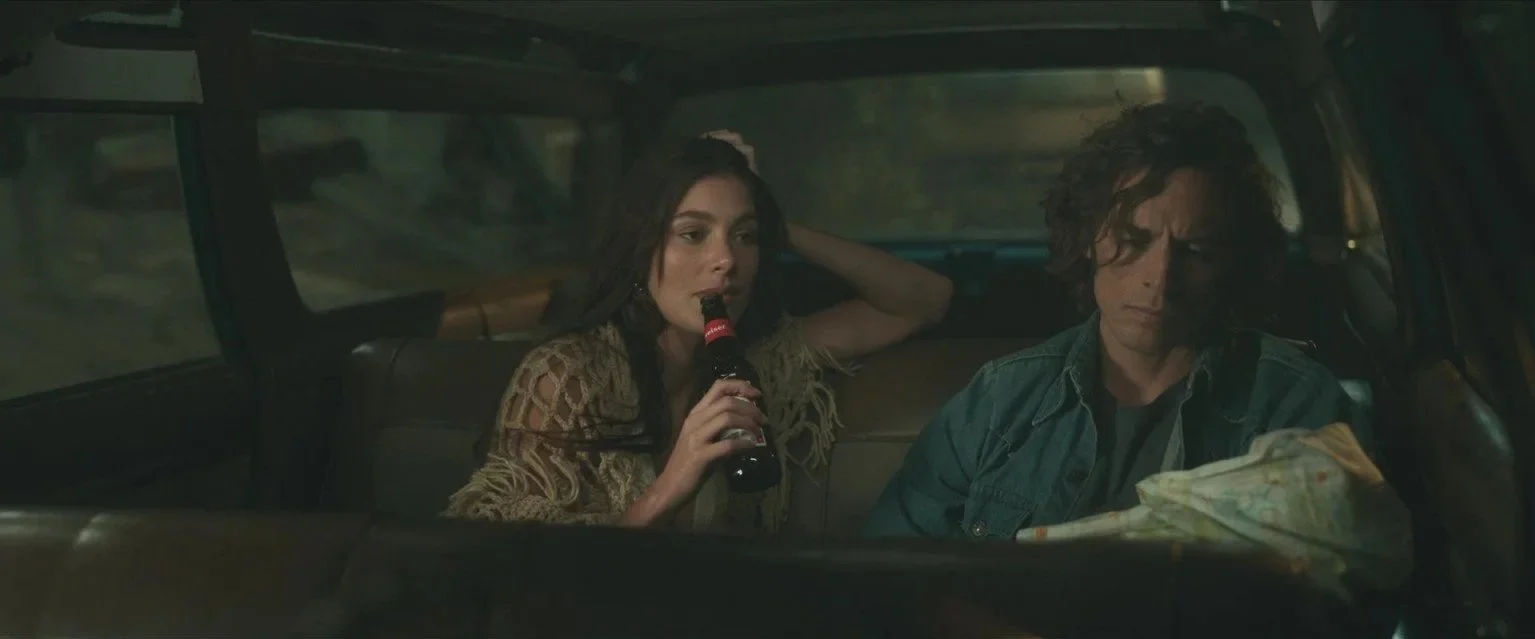 Two people sitting in the back of a vehicle, the woman is drinking a soda and has her hand in her hair, and the man is looking at a map.