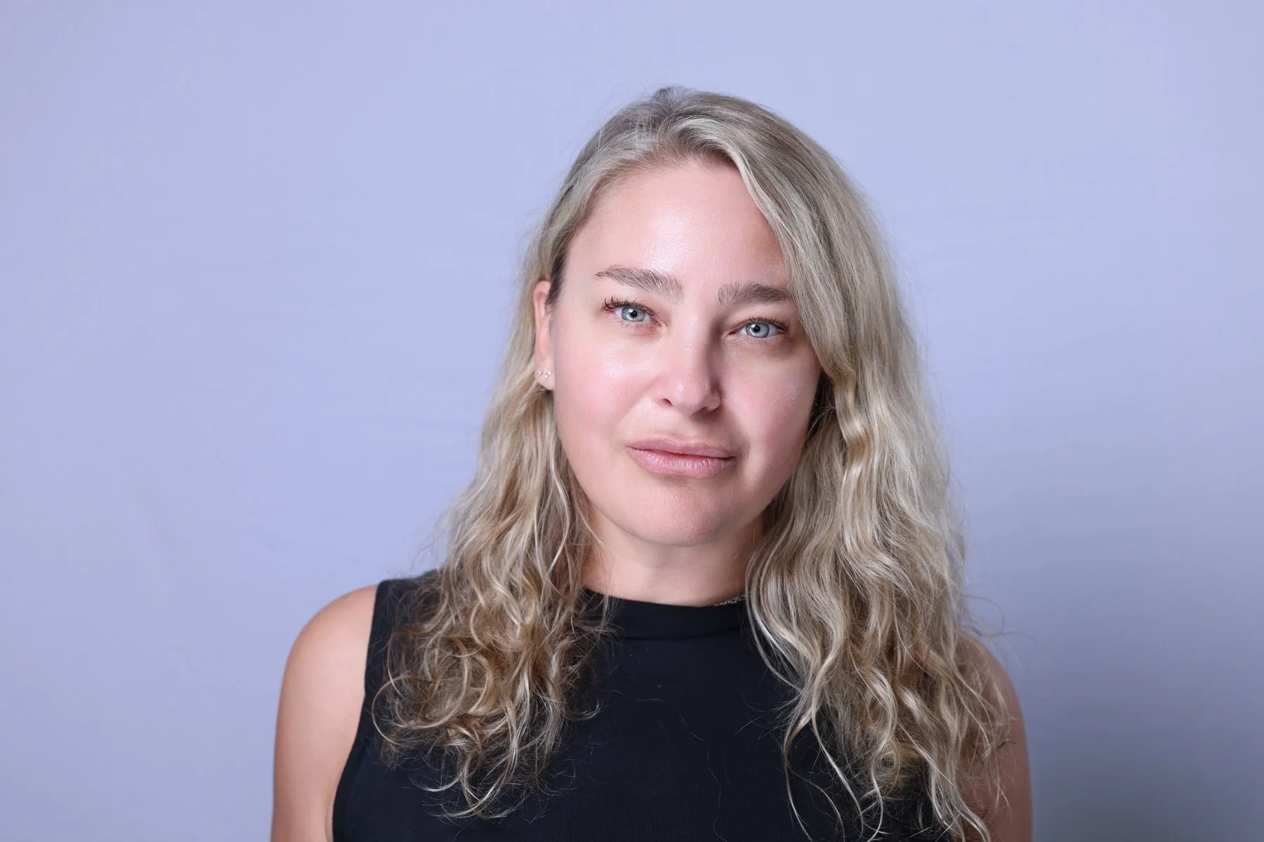 A woman with blonde, curly hair wearing a black sleeveless top standing against a light purple background.