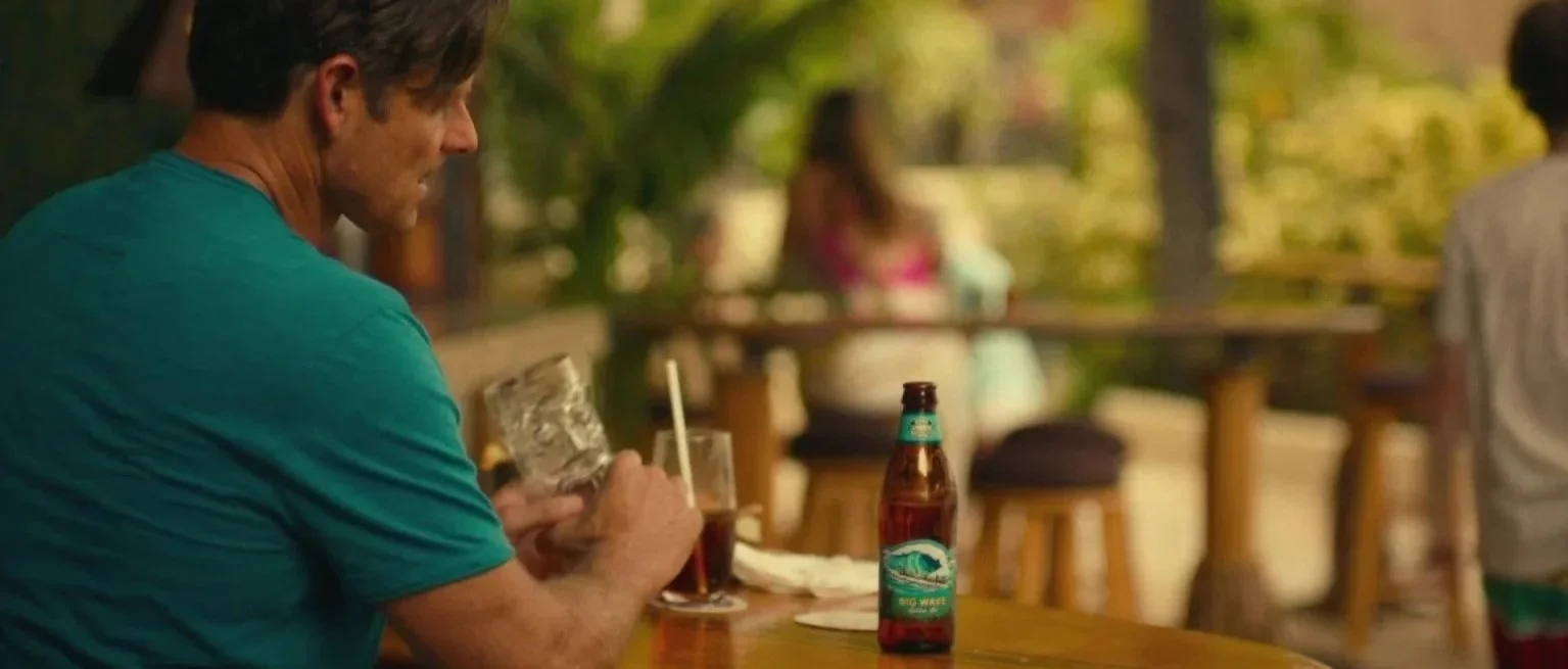 A man sitting at a wooden table outdoors on a patio, holding a glass of dark beer, with a bottle of beer on the table beside him, and another person blurred in the background.