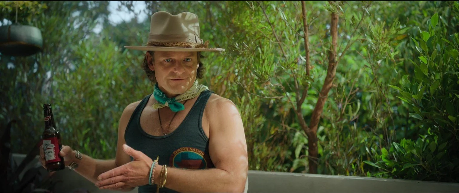 A man wearing a tan floppy hat, a teal neckerchief, and a sleeveless black tank top with a logo, standing outdoors with green foliage in the background. He is holding a beer bottle.