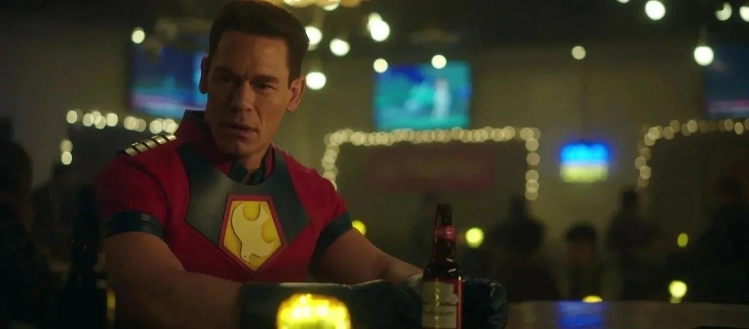 A man dressed as Superman with a superhero emblem on his chest is sitting at a bar, looking surprised or concerned, with a beer bottle and glasses on the counter, and blurred TV screens and lights in the background.