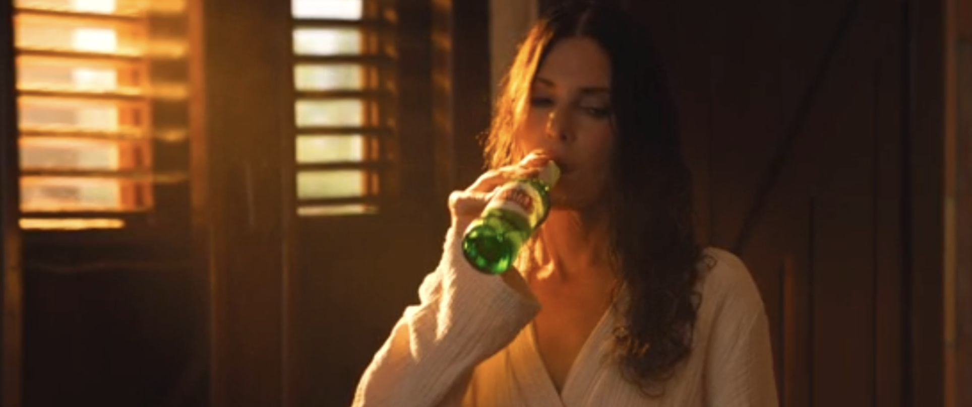 A woman with dark hair drinking from a green glass bottle in a dimly lit room with wooden blinds.