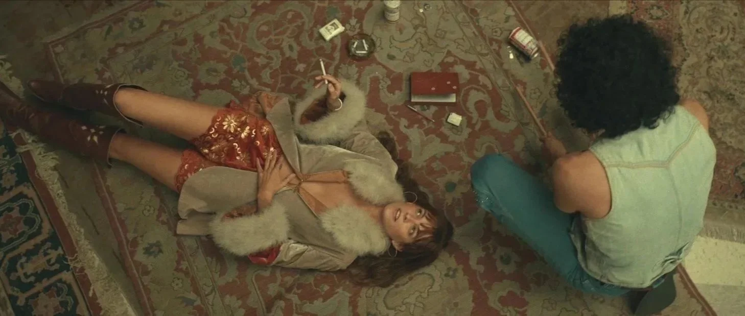 A woman lying on a rug looking up with a man sitting cross-legged nearby, both in casual clothing, with various objects scattered around them.