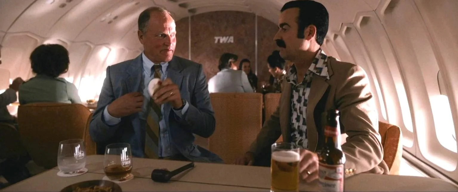 Two men having a conversation on an airplane. One man wearing a gray suit and the other in a tan jacket with a beer and a bottle on the table.