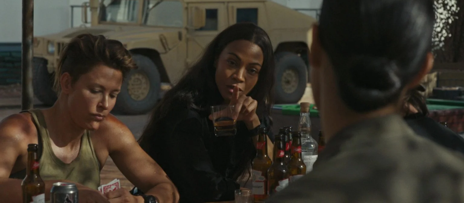 Three women sitting at a table with drinks, playing cards, and bottles of beer in a casual outdoor setting.