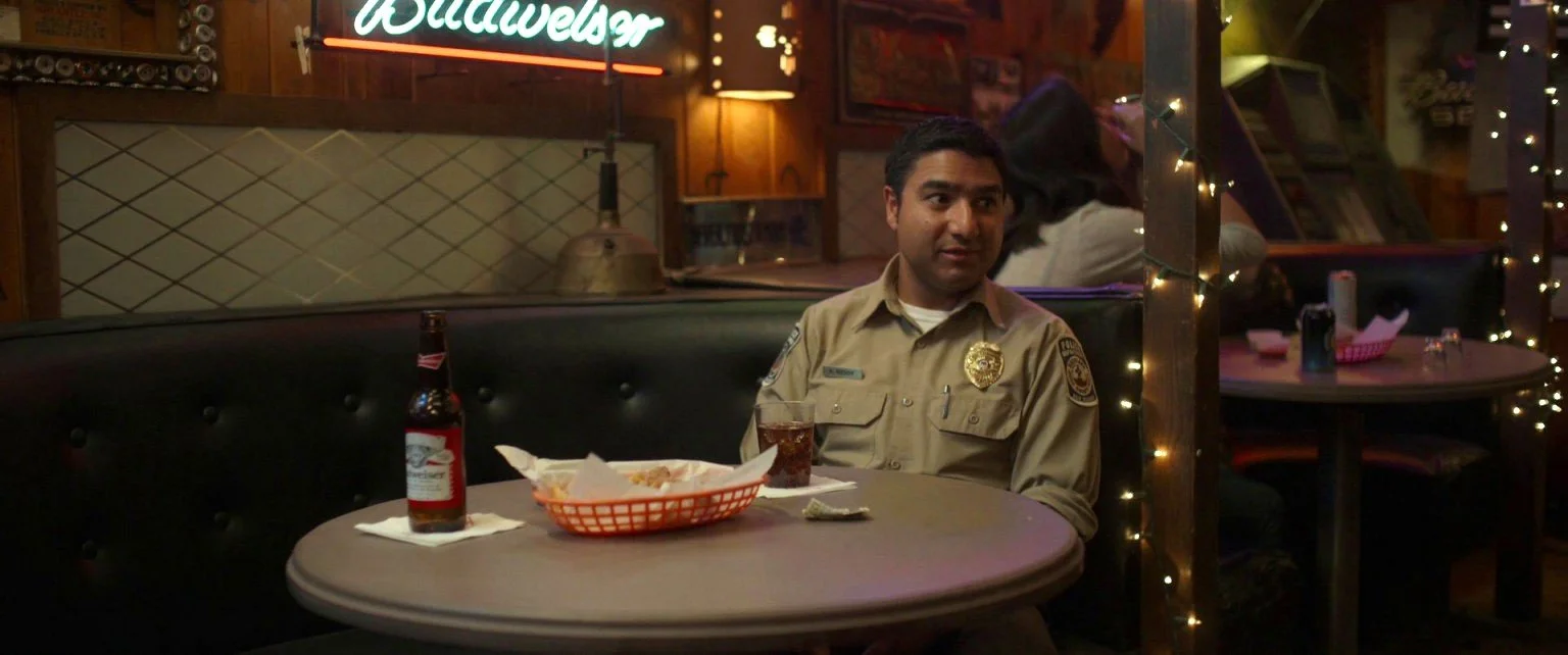 A man in a sheriff's uniform sitting at a table in a restaurant or bar, with a beer bottle, a glass of soda, and a basket of food on the table, decorated with fairy lights.