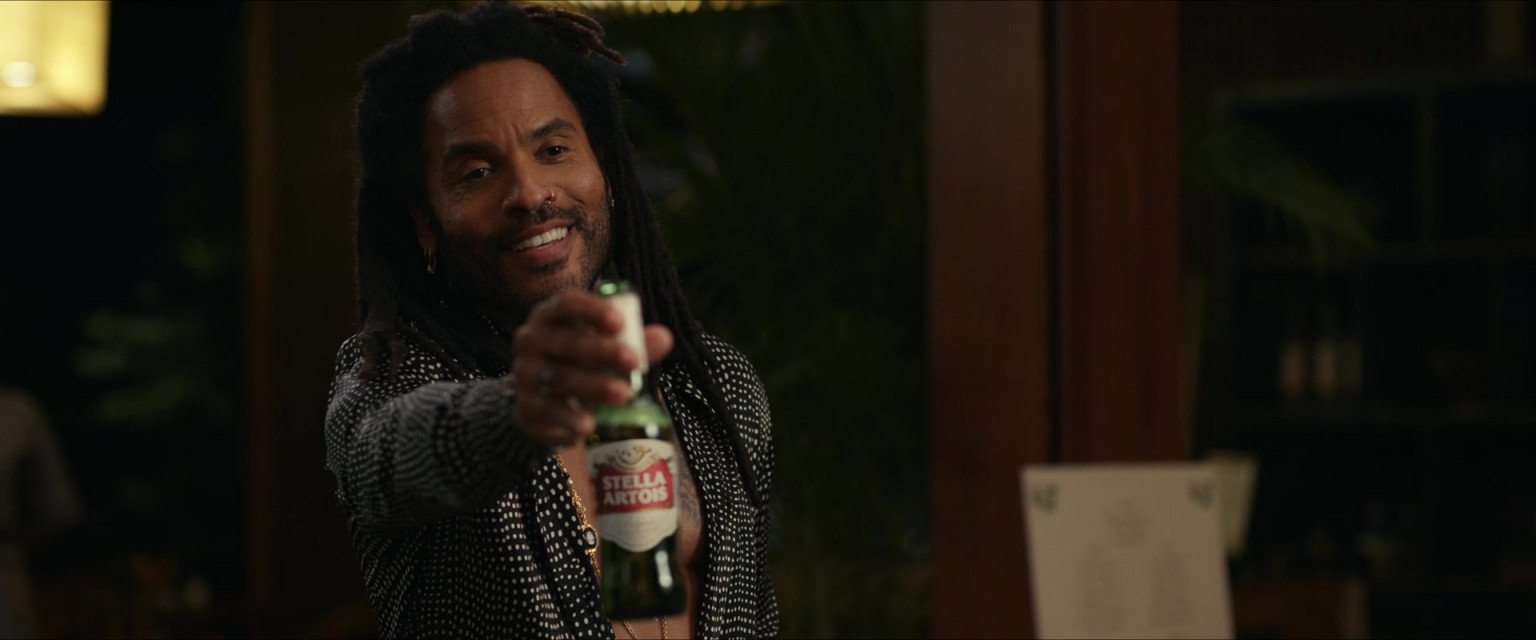 A man with dark skin, dreadlocks, and a goatee, smiling and holding a Stella Artois beer bottle toward the camera, in a dimly lit room with a dark background.