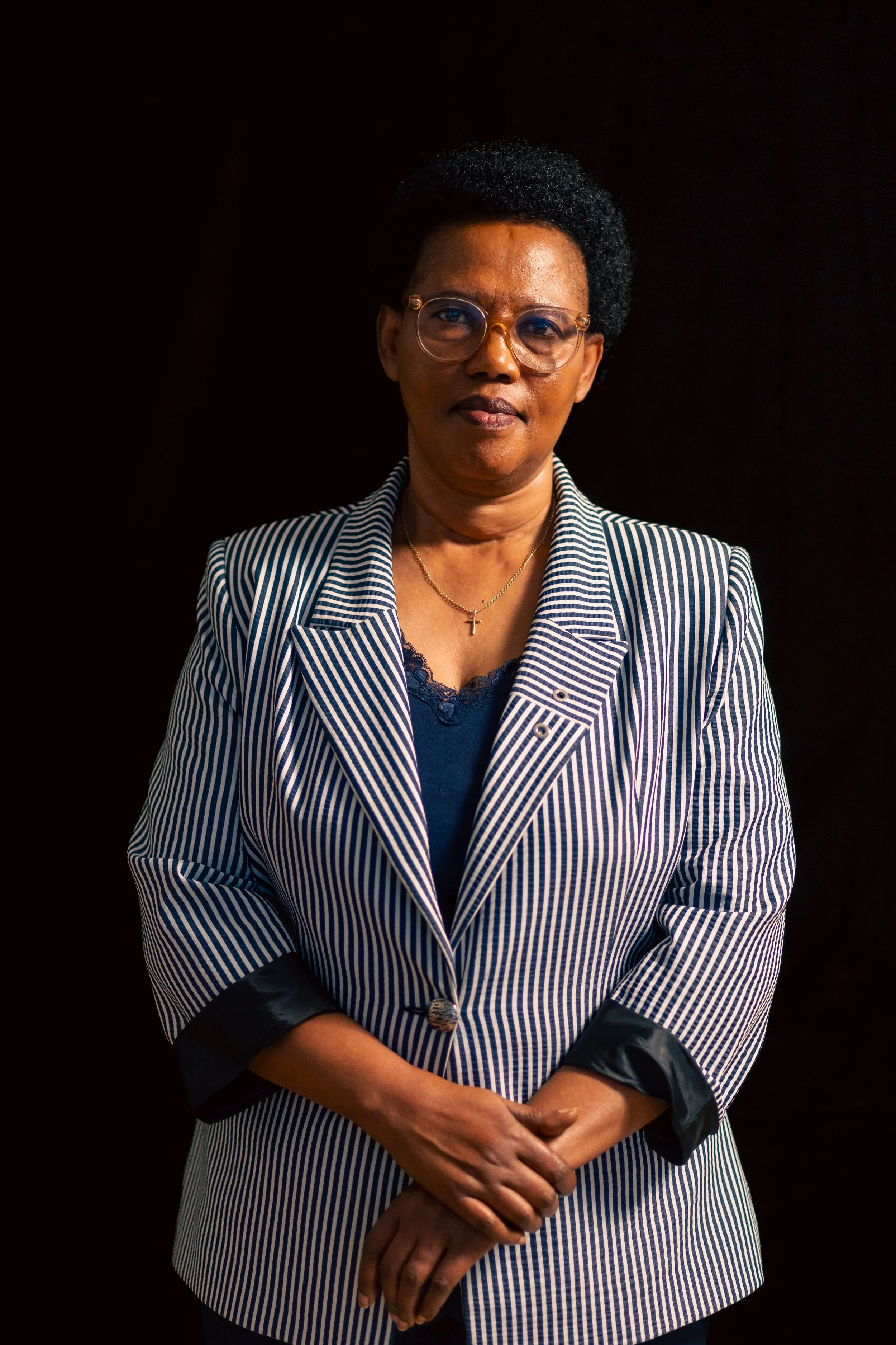 A woman with short curly black hair, glasses, and wearing a striped blazer with a cross necklace, standing against a black background.