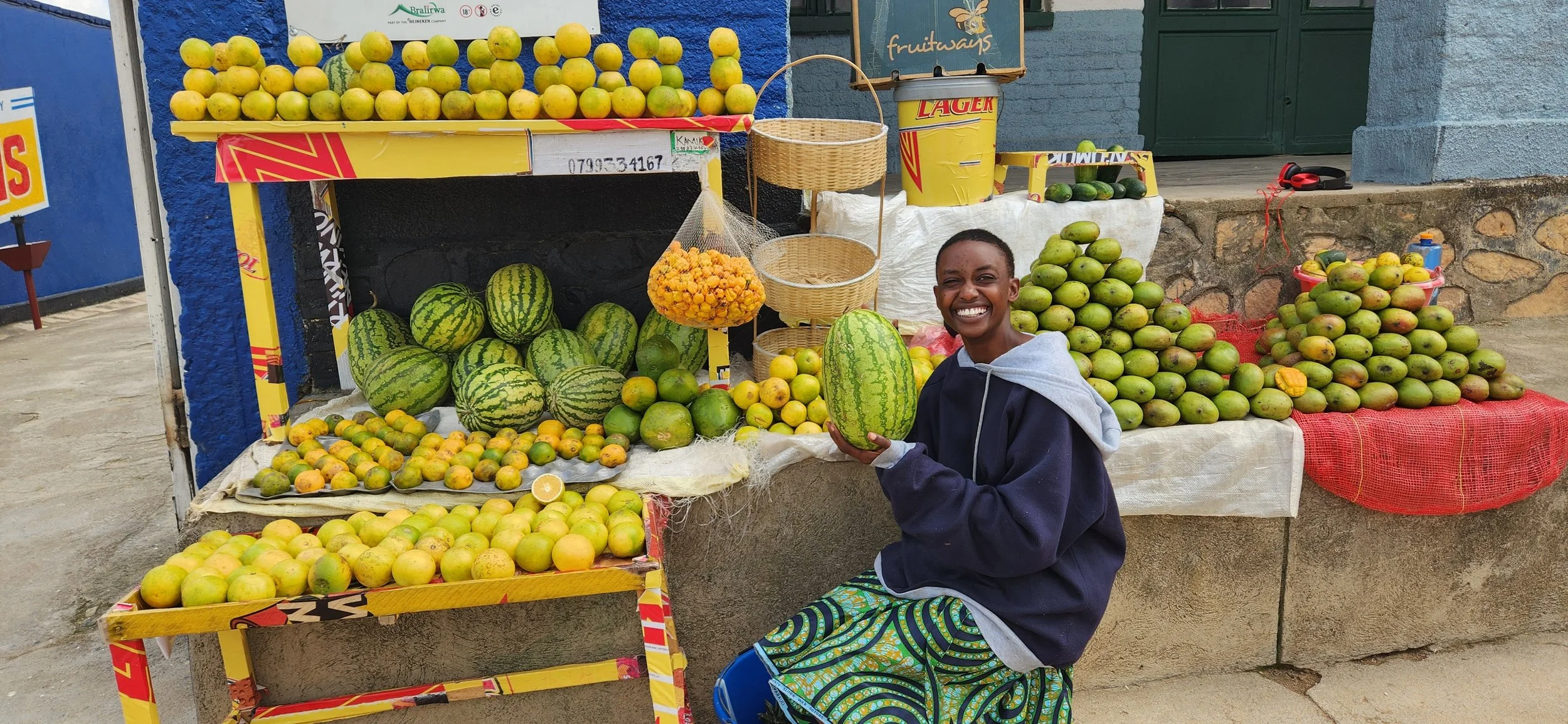 A woman sitting in front of a fruit stand with watermelons, papayas, and other fruits on display. She is smiling and holding a small watermelon.