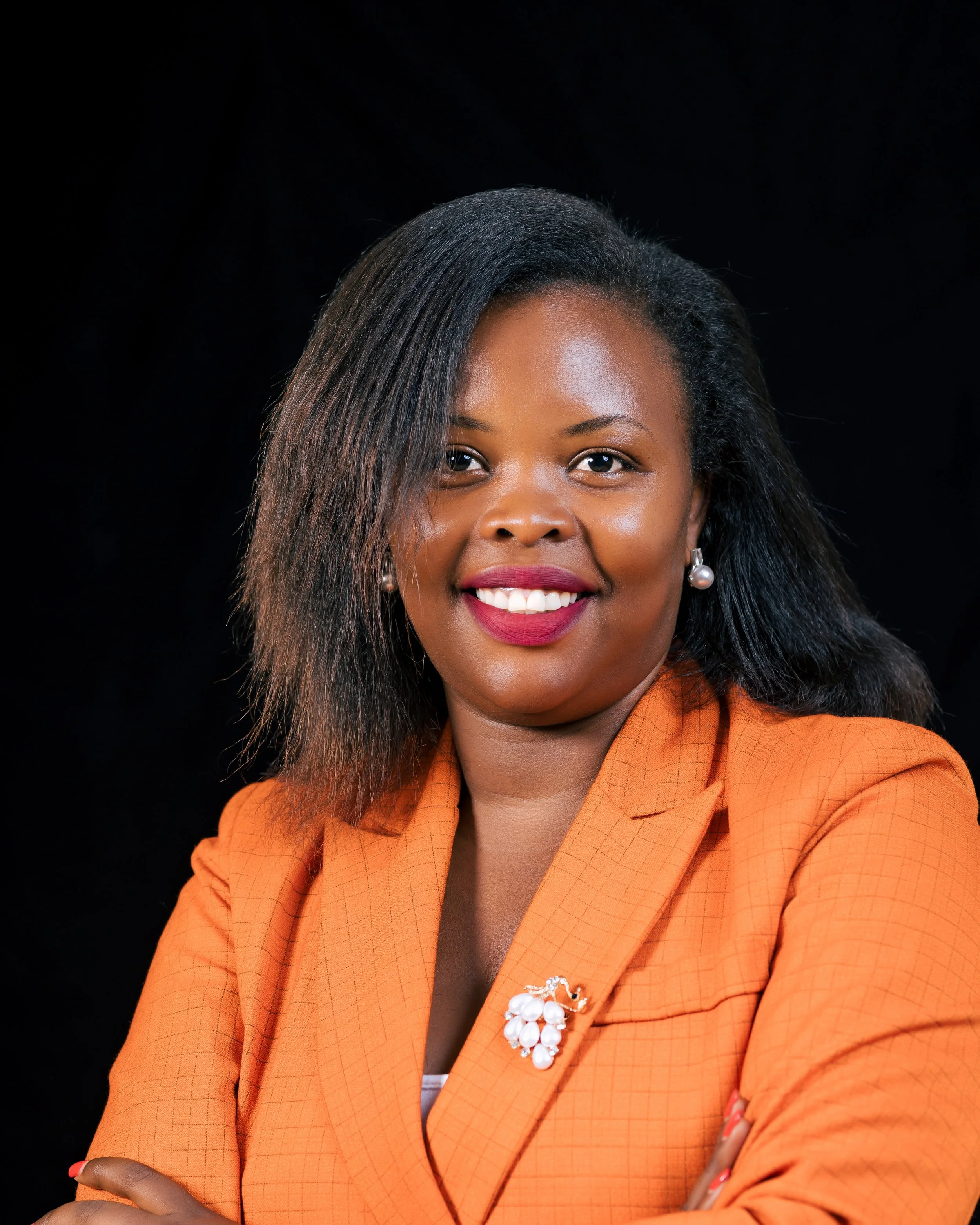 Close-up of a woman with dark skin wearing an orange blazer, pearl earrings, and red lipstick, smiling against a black background.