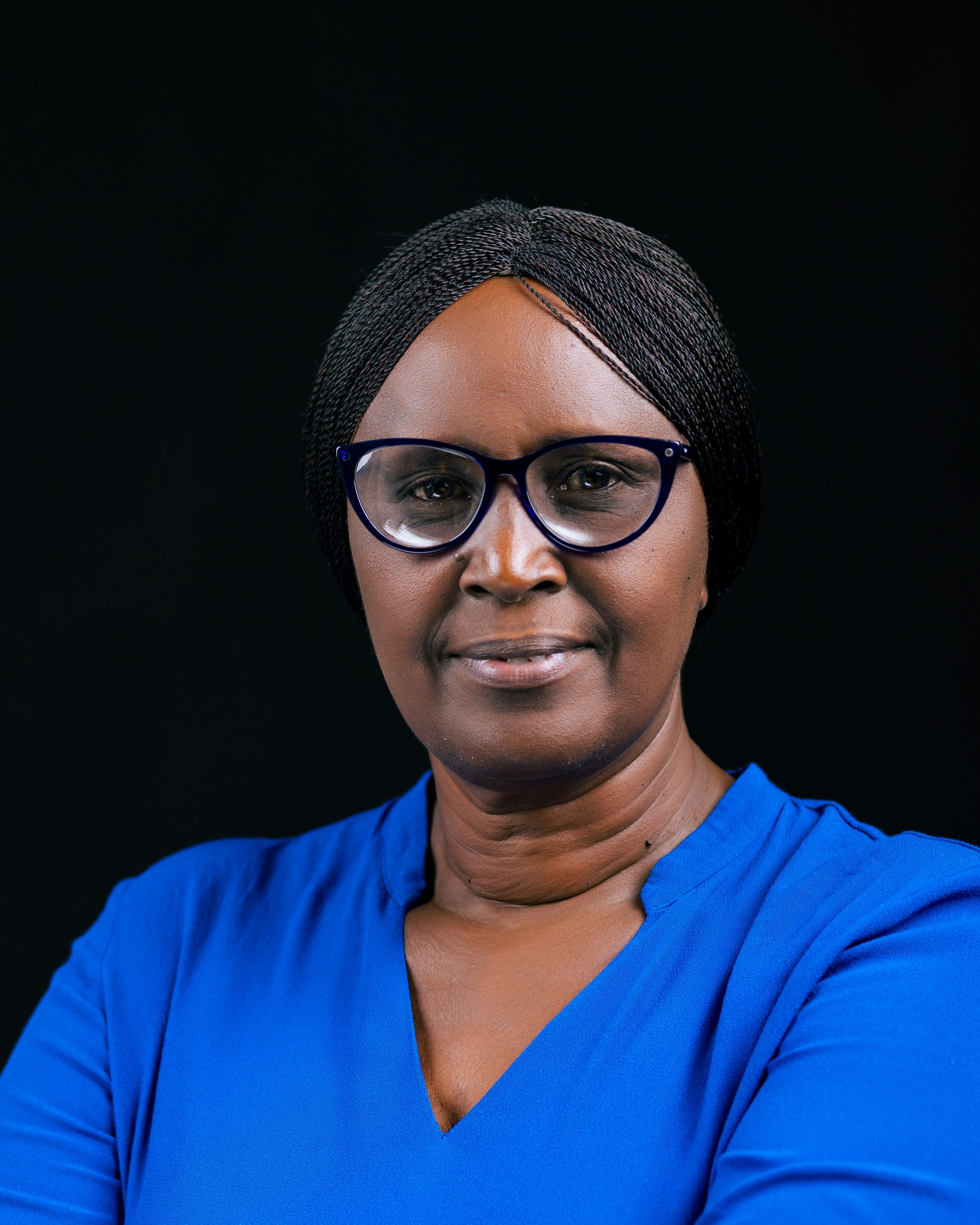 Close-up portrait of a woman with medium dark skin, wearing black-framed glasses and a blue top, against a black background.