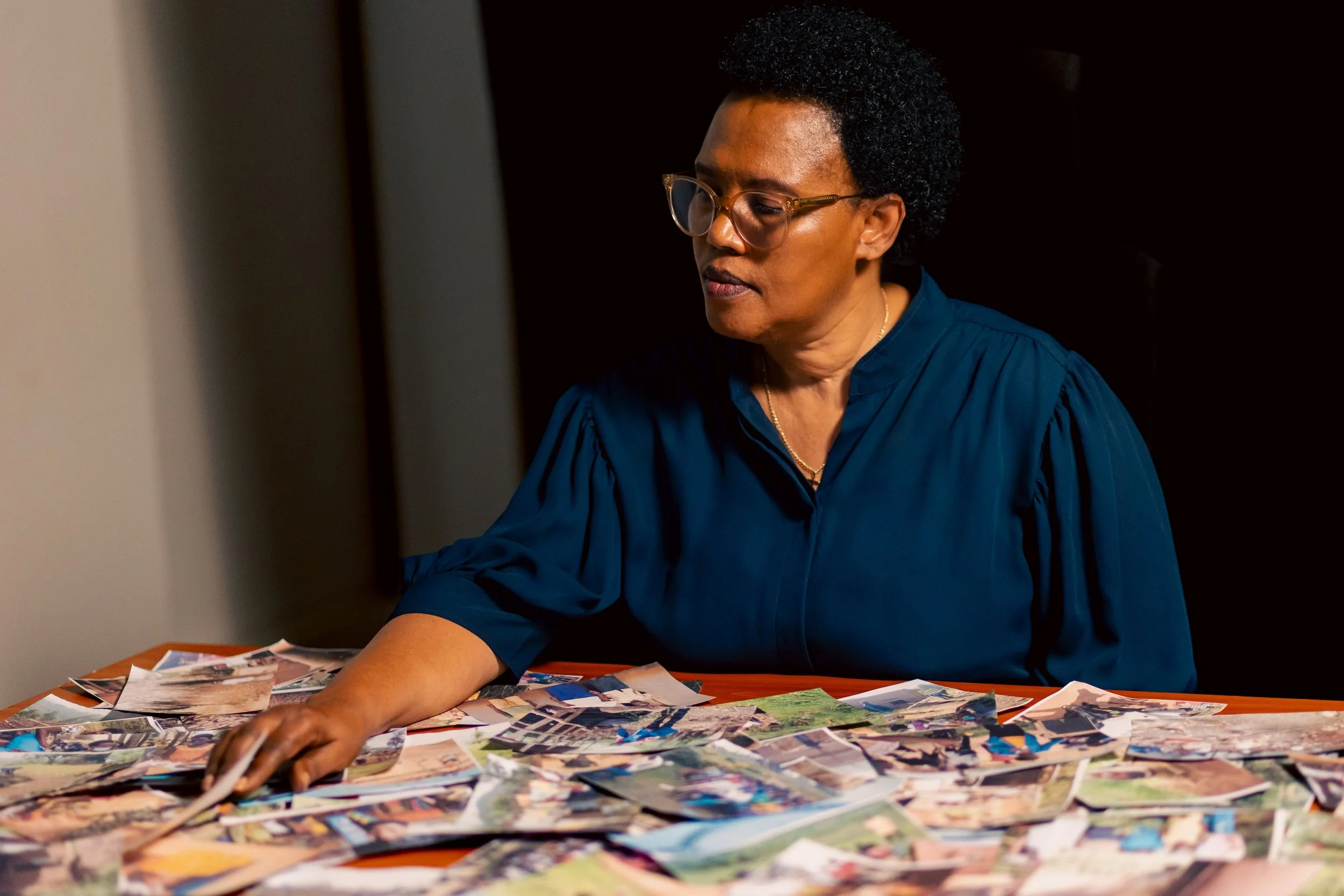 A woman with short curly hair, glasses, and wearing a blue shirt, organizing and sorting through a large collection of photographs on a table.