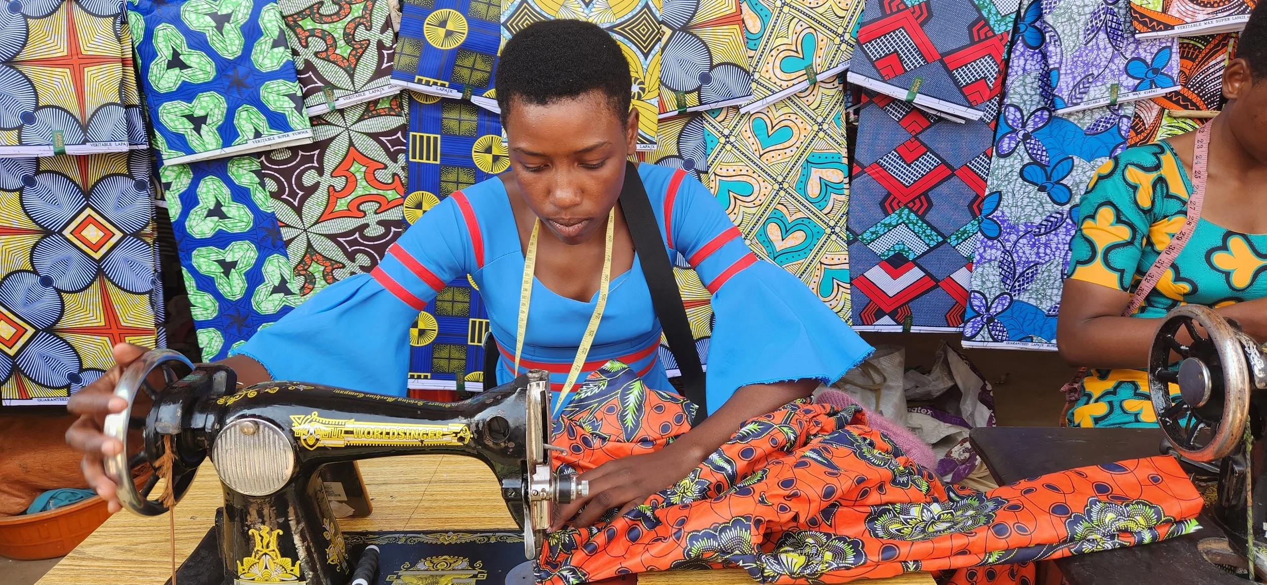 A woman in a blue dress with red and yellow accents is sewing fabric on a vintage black sewing machine labeled "WORLD SINGER" at a colorful fabric stall. Behind her are vibrant, patterned fabrics hanging, featuring various geometric and floral designs in bright colors.