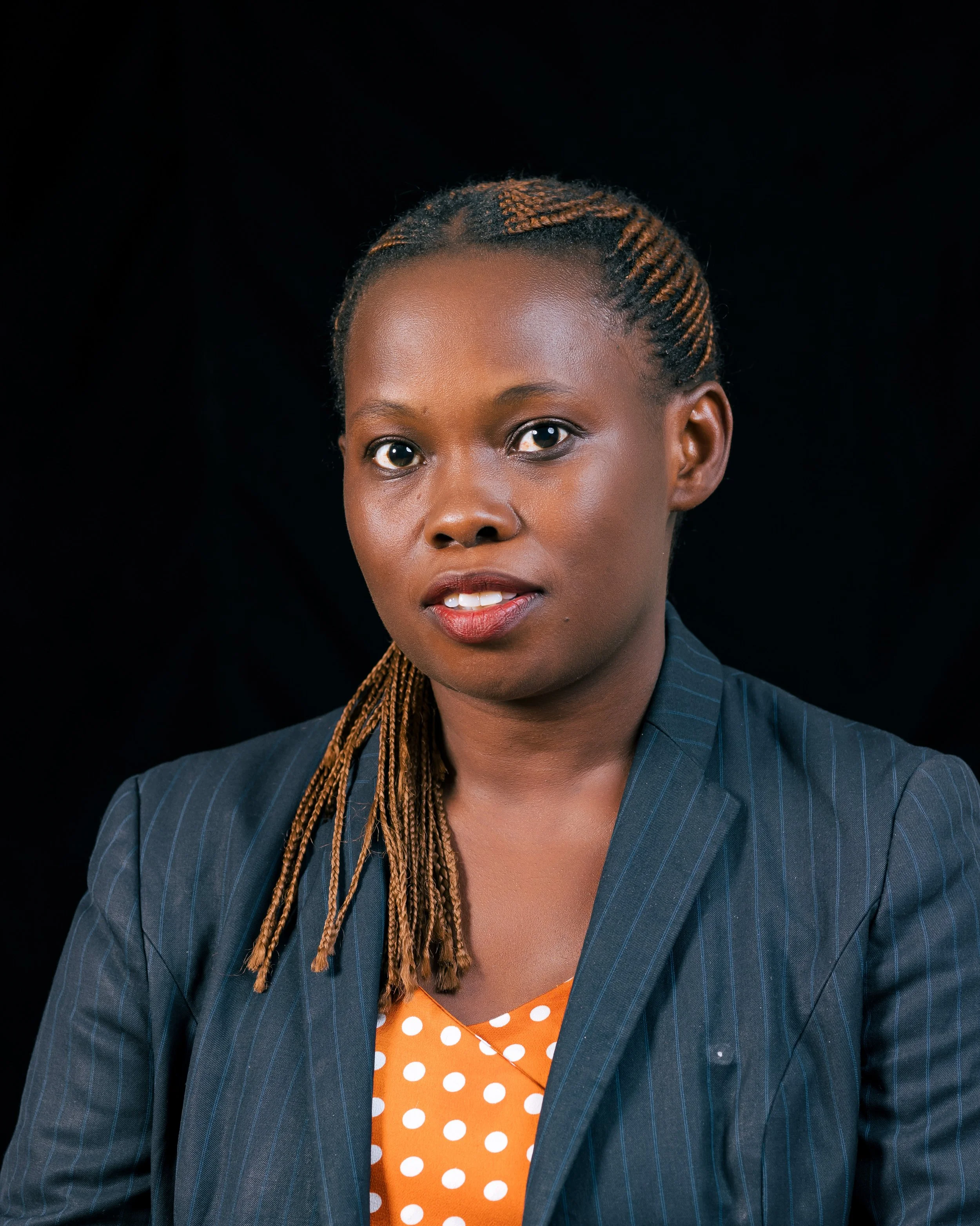 Close-up portrait of a woman with braided hair, wearing a blue pinstripe blazer over an orange polka dot blouse, against a black background.