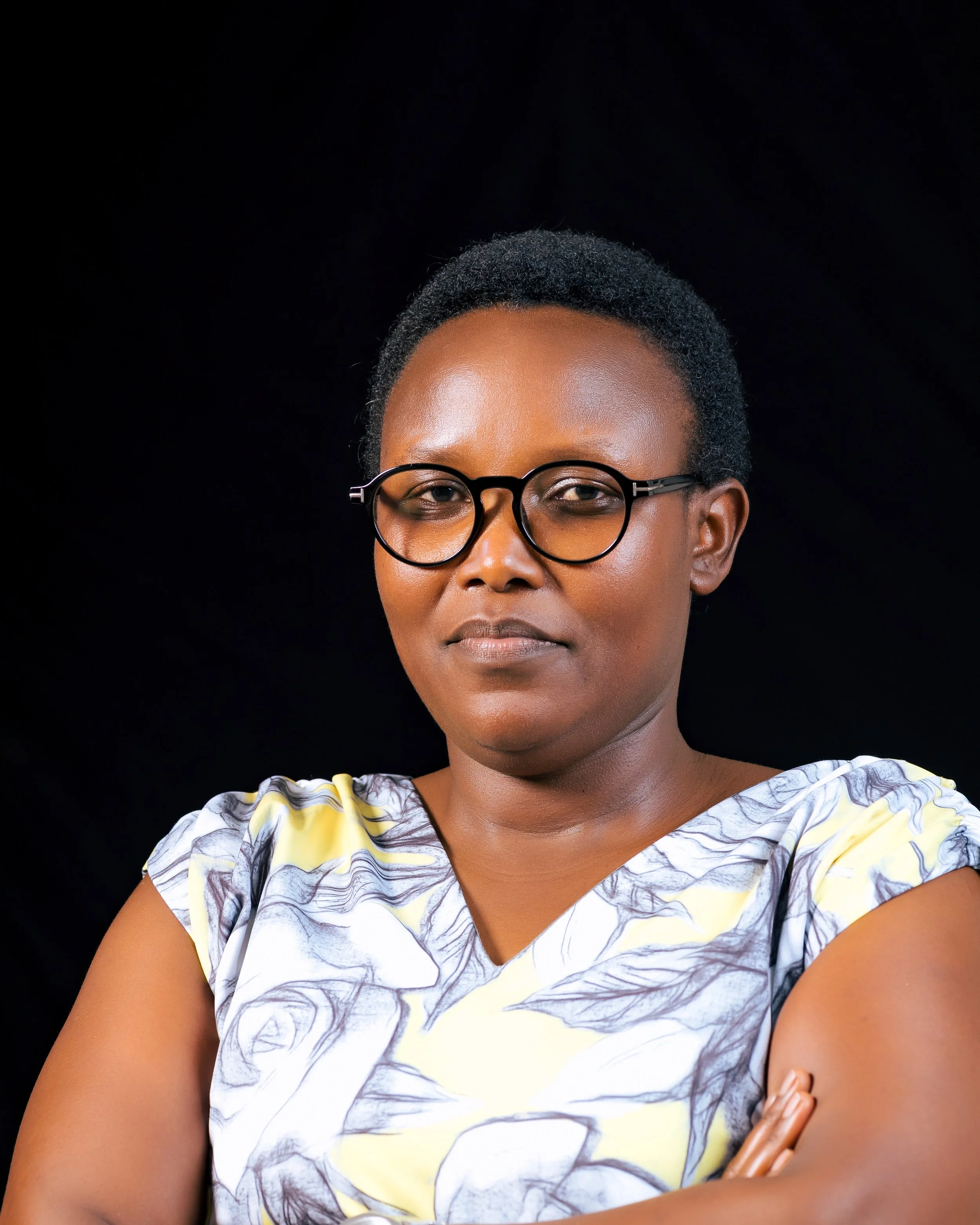 Close-up of a woman with dark skin, short textured hair, and round glasses, wearing a yellow and gray patterned top, against a black background.