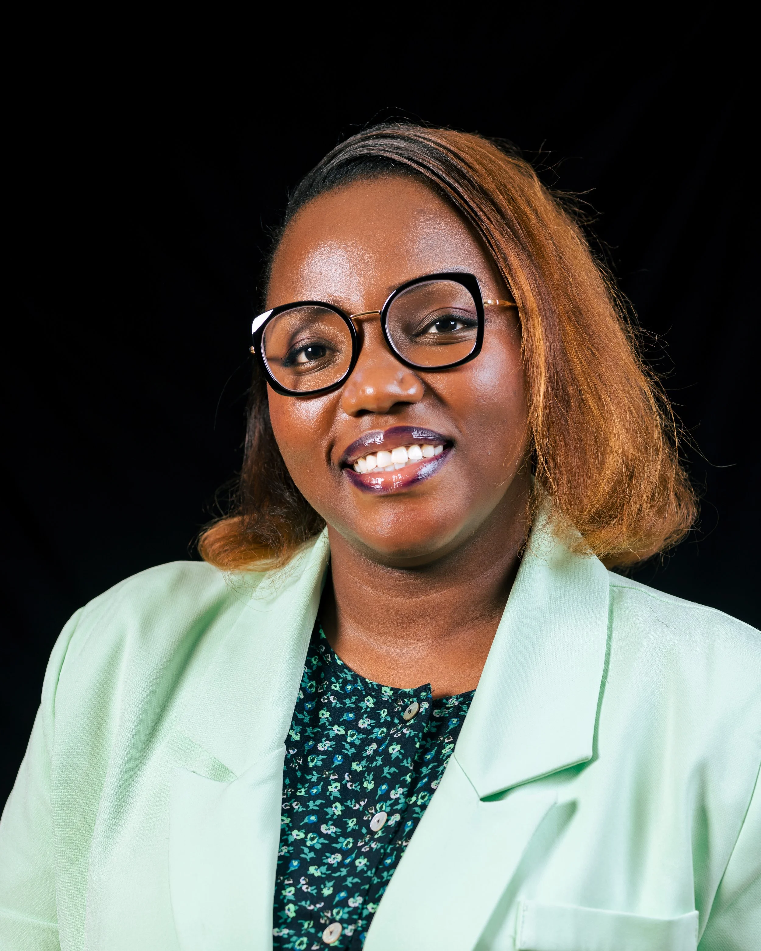 Close-up of a woman with brown skin wearing large black rim glasses, smiling, and dressed in a light green blazer over a dark floral top, against a black background.