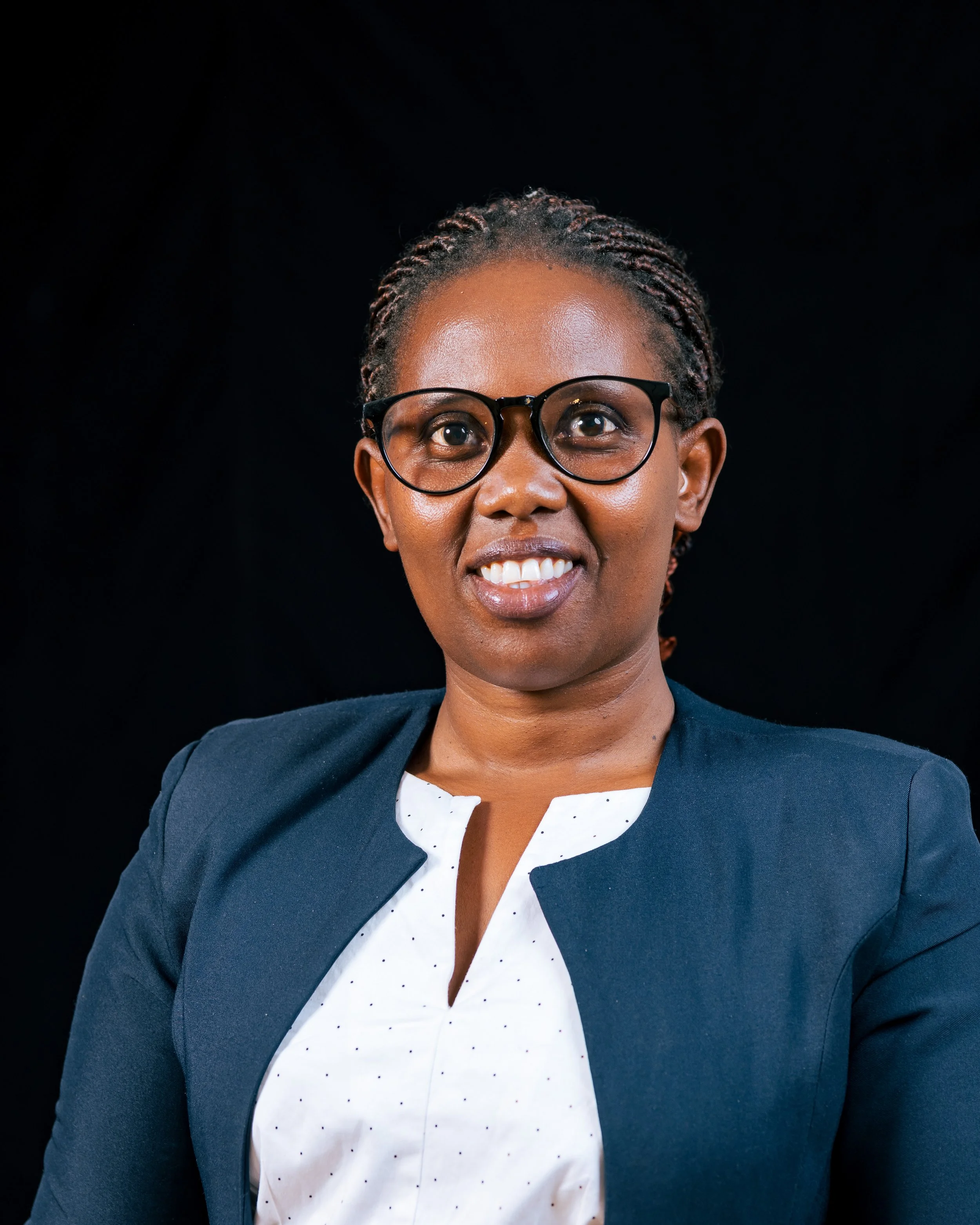 A smiling woman with dark skin, glasses, and braided hair, dressed in a professional outfit, standing against a black background.