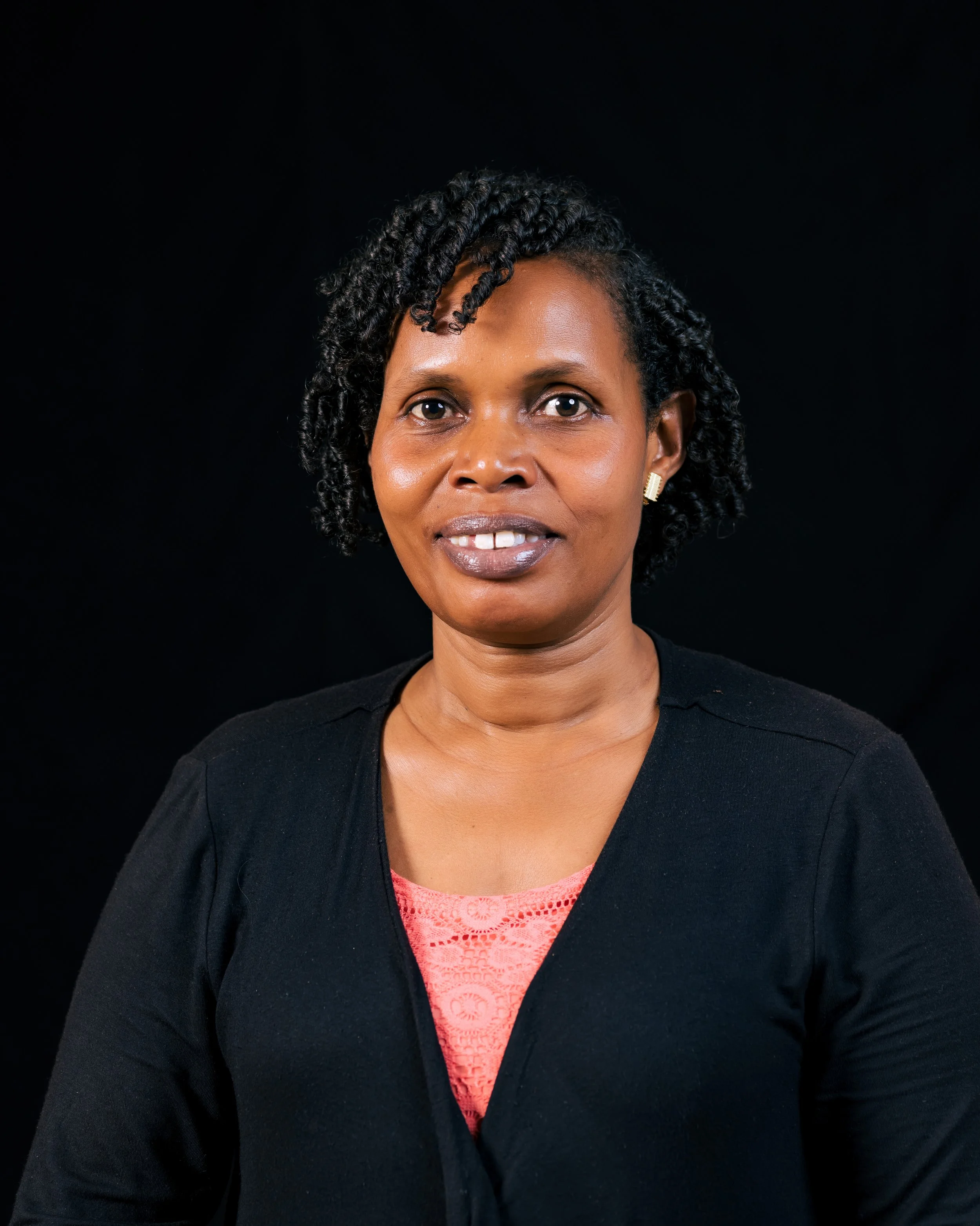 Close-up portrait of a smiling woman with short, curly hair, wearing a black cardigan over a coral lace top, set against a black background.