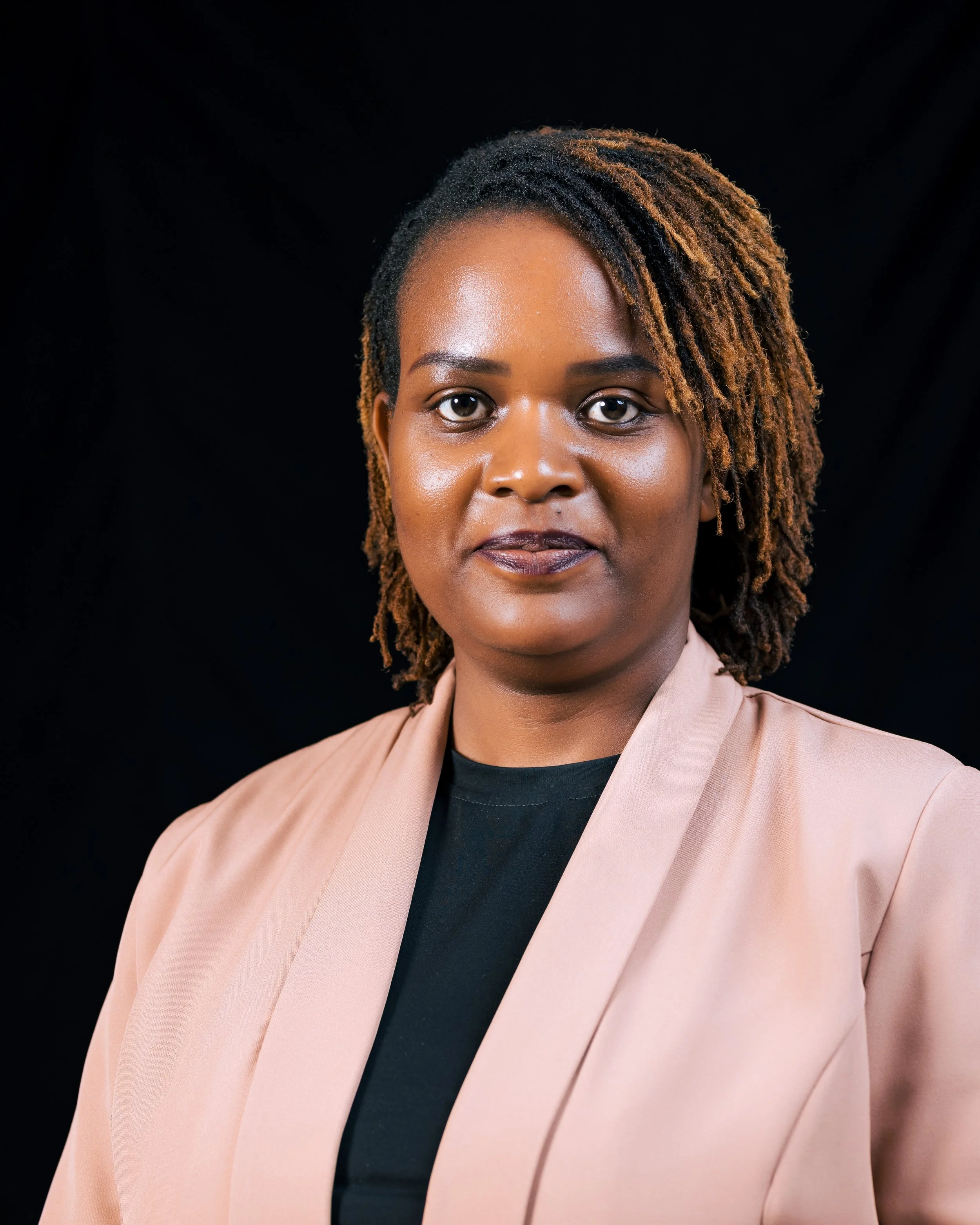 A portrait of a woman with dark skin, dreadlocks, and wearing a beige blazer over a black top, against a black background.