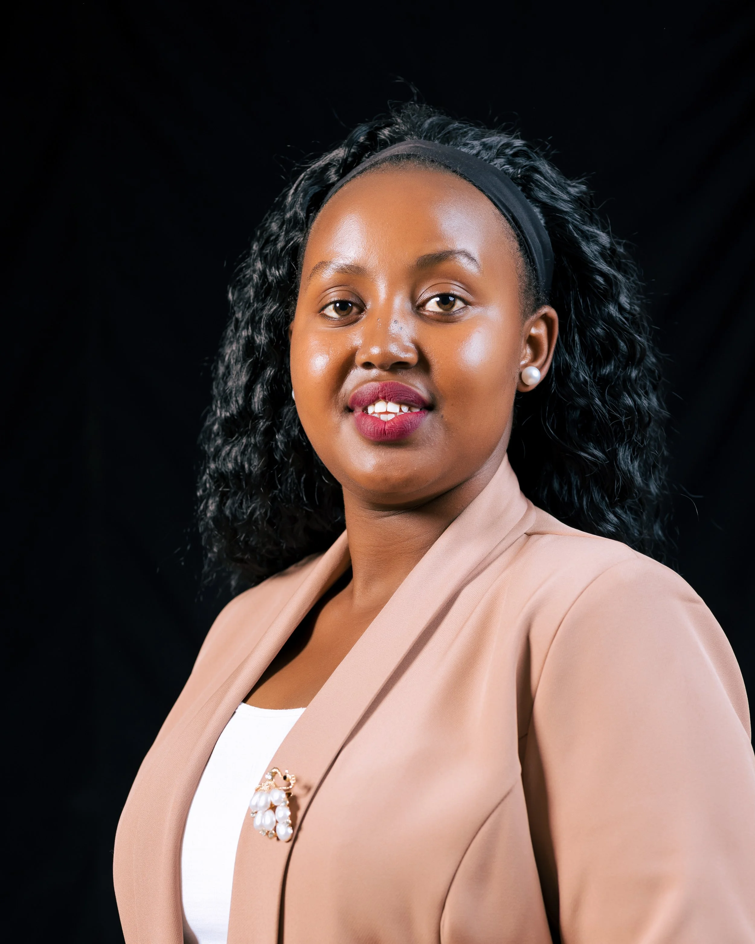 A woman with curly black hair, wearing a black headband, pearl earrings, a beige blazer, and a white top with a pearl brooch, posing against a black background.
