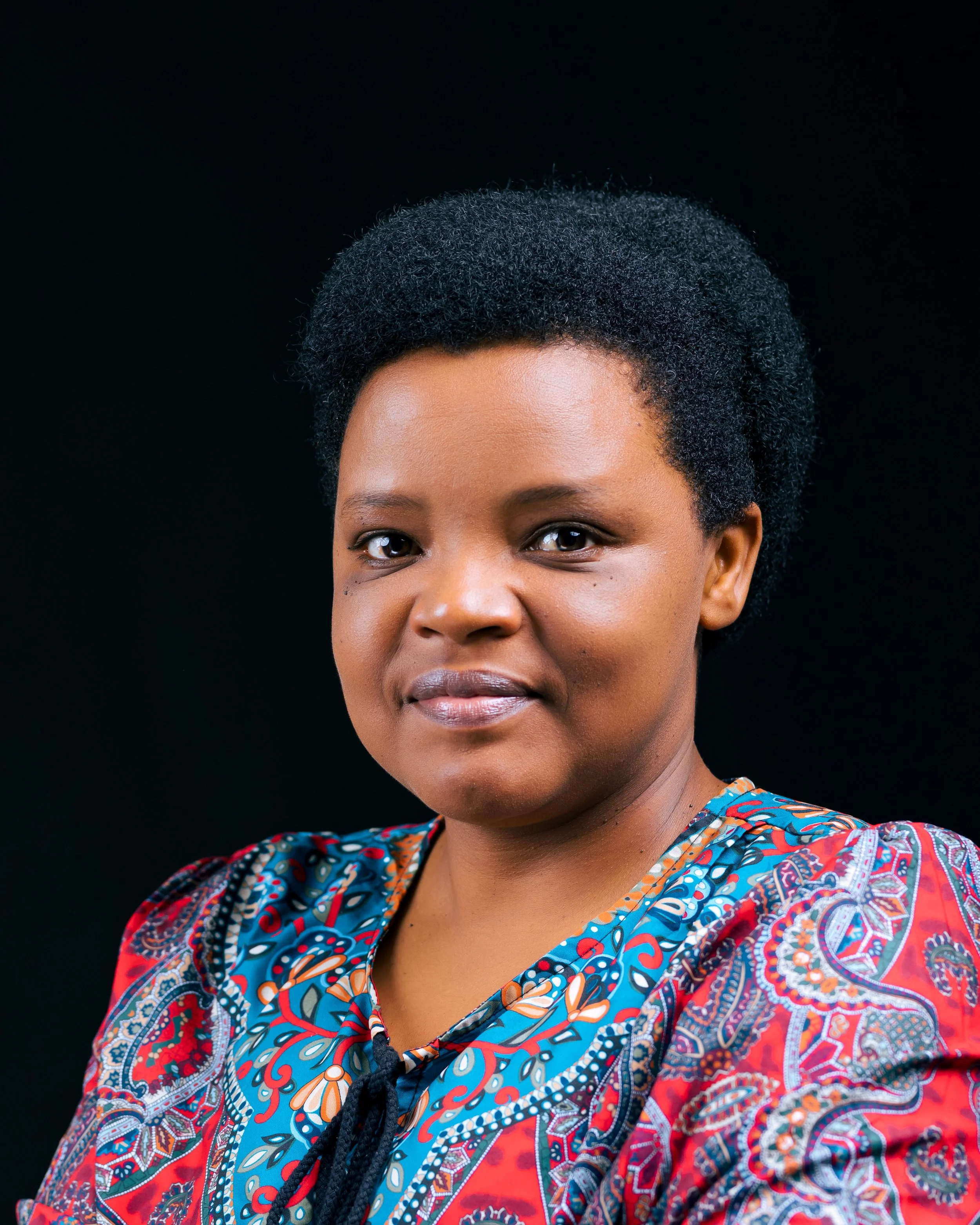 A close-up of a woman with short, curly black hair, wearing a colorful patterned shirt, against a black background.