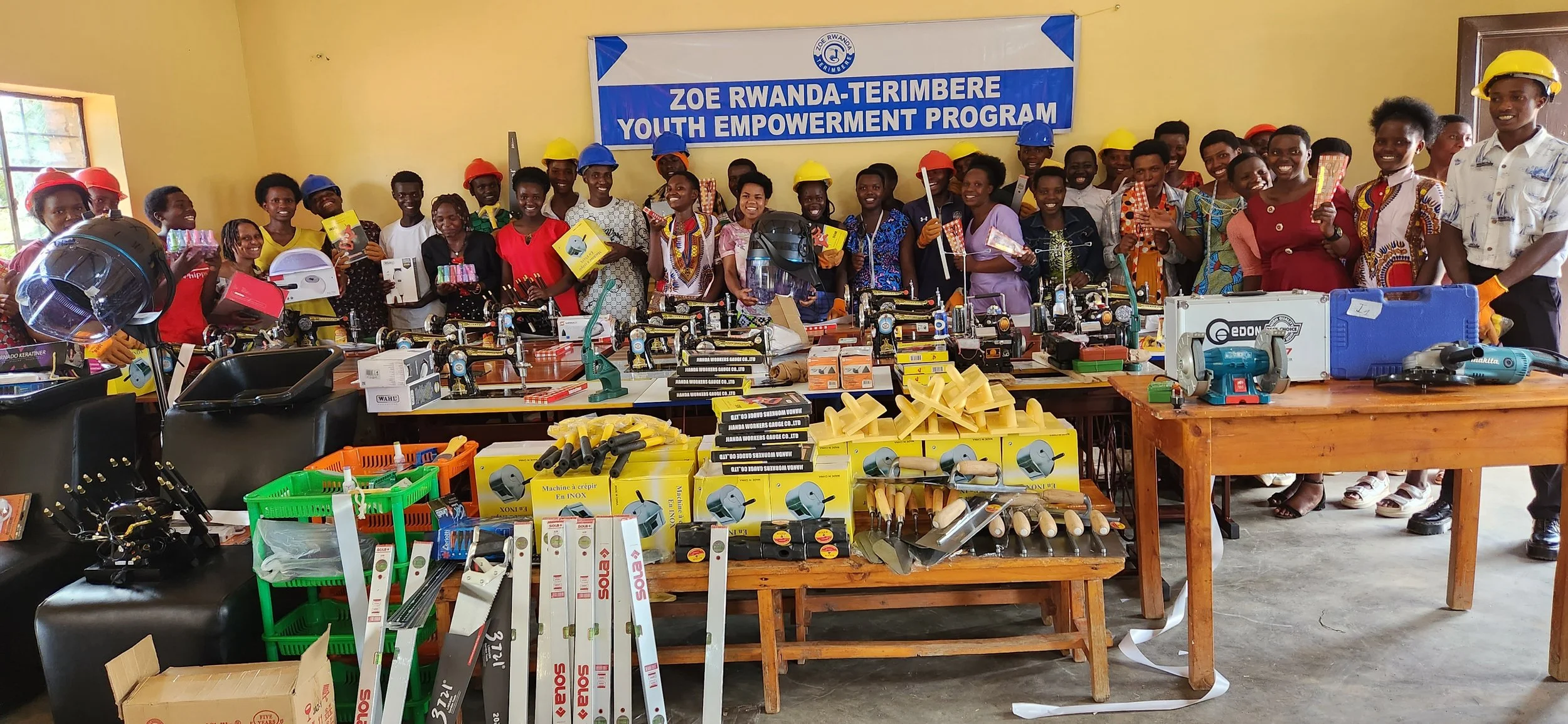Group of young people at an empowerment program in Rwanda, wearing colorful helmets, standing behind tables filled with tools and hardware equipment, with a banner that reads 'Zoe Rwanda-Terimbere Youth Empowerment Program'.
