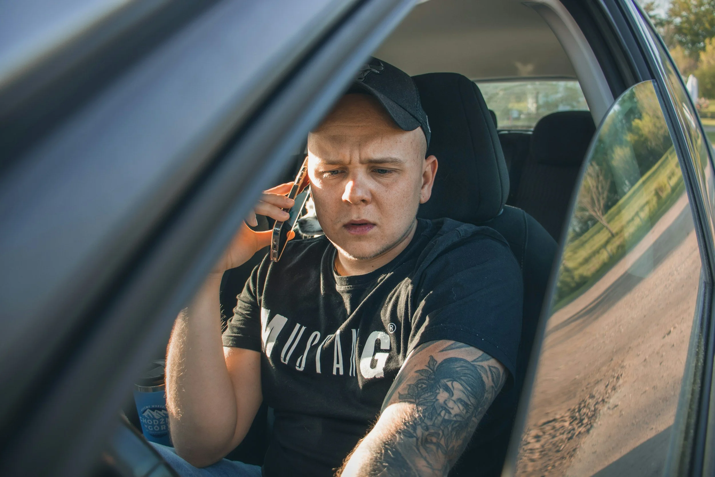 Young man with tattoos on his arm, wearing a black T-shirt and baseball cap, talking on the phone inside a car, sitting in the driver's seat.